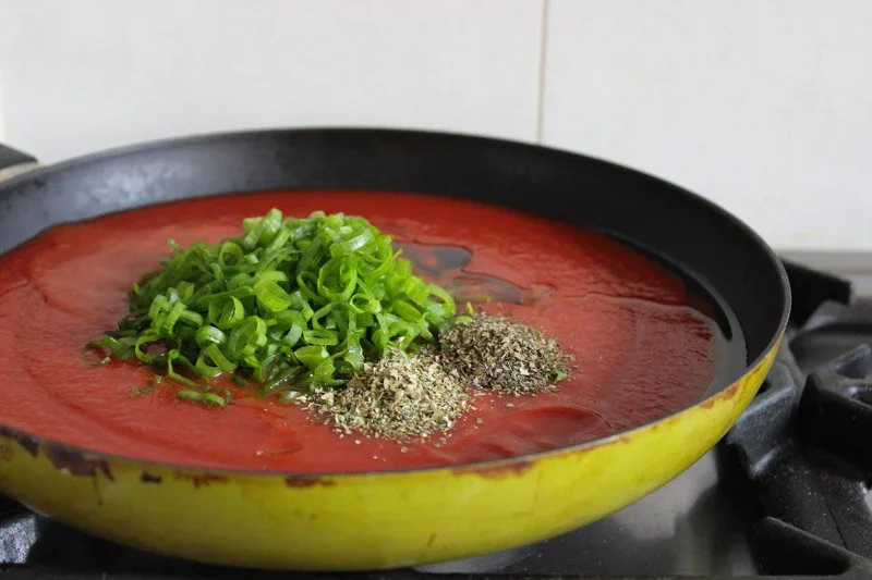 A side-angle view of a frying pan filled with smooth tomato puree, topped with a pile of chopped green spring onion tops and dried herbs.