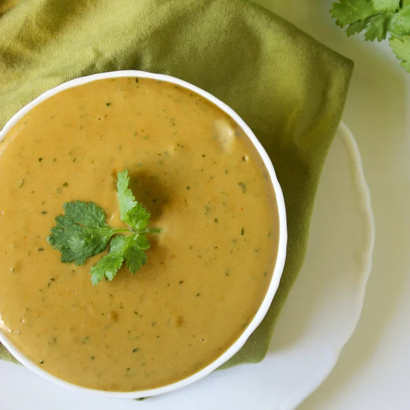 A top-down view of a white bowl filled with creamy low FODMAP satay sauce, garnished with a fresh coriander leaf on a green napkin.