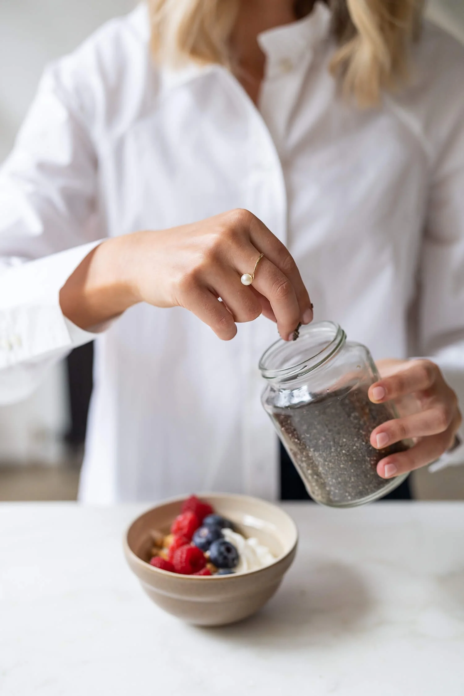Person seasoning yogurt bowl with berries, wearing a white shirt and a pearl ring.
