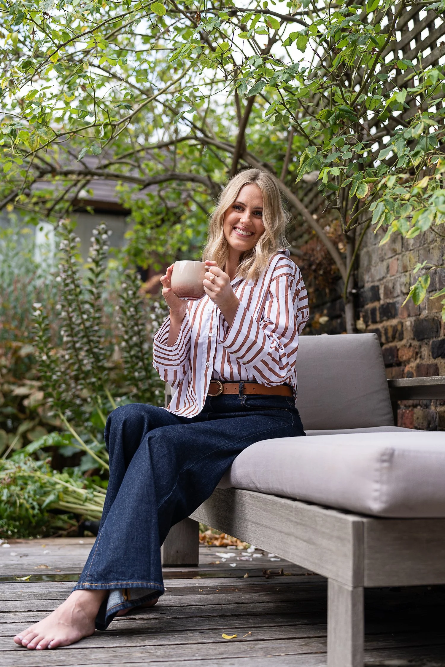 A woman sitting on a outdoor wooden bench, holding a mug, smiling, wearing a striped shirt and blue jeans, surrounded by greenery and a brick wall.