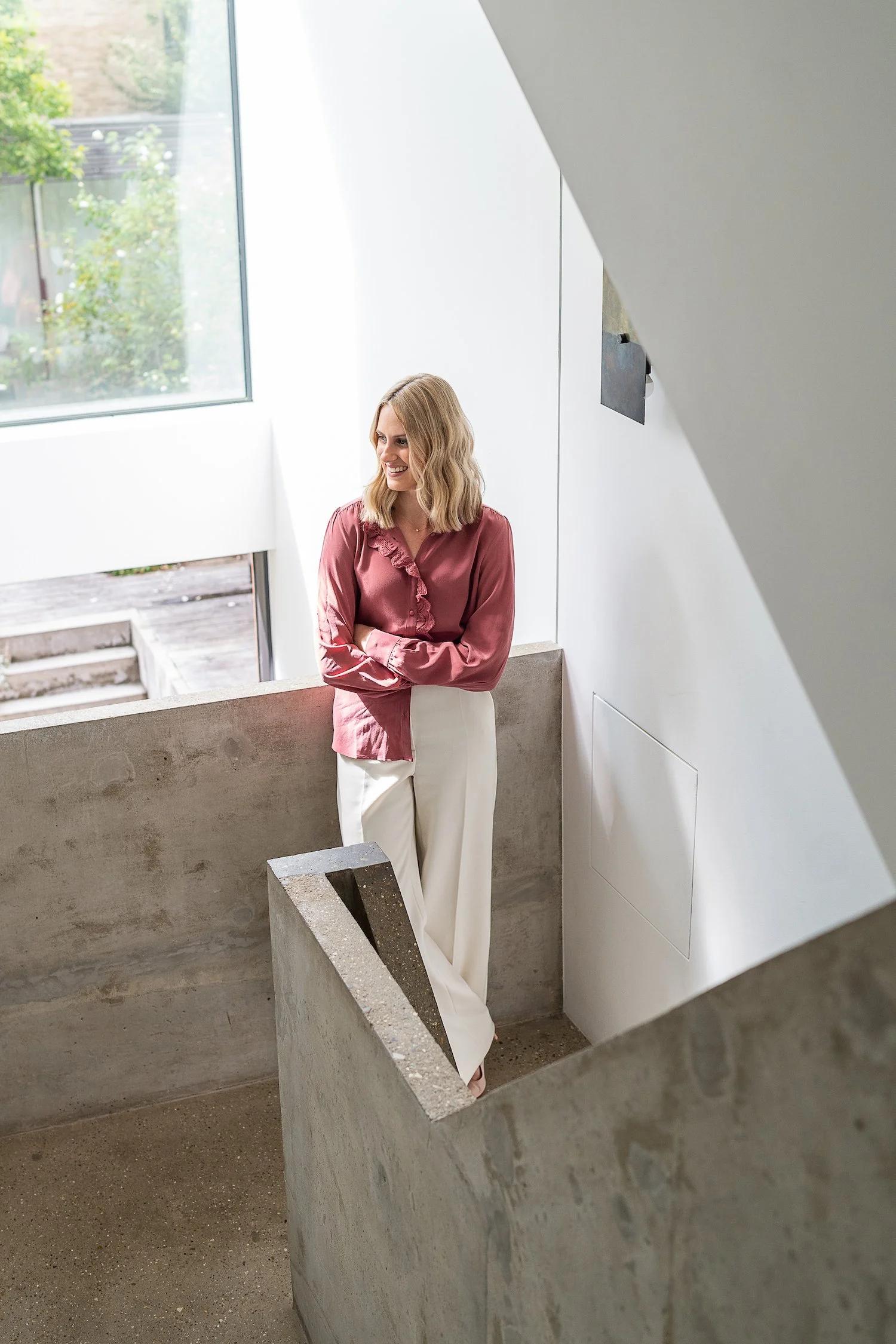 A woman with blonde hair wearing a maroon blouse and cream-colored pants standing in a modern, minimalistic stairwell with concrete walls and large windows.