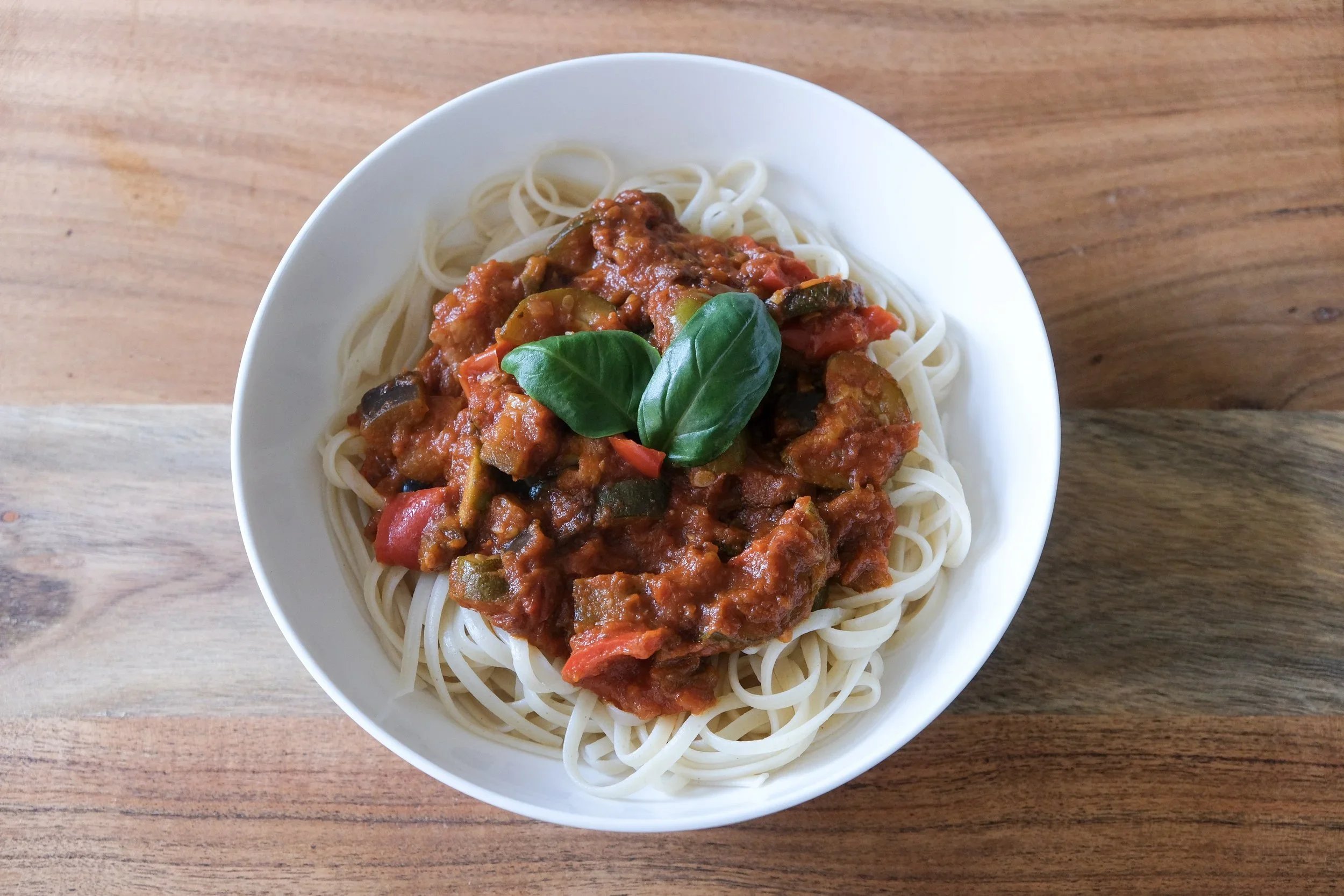 A white bowl of long pasta noodles topped with a rich tomato vegetable sauce containing roasted zucchini, eggplant, bell peppers, and fresh basil leaves.