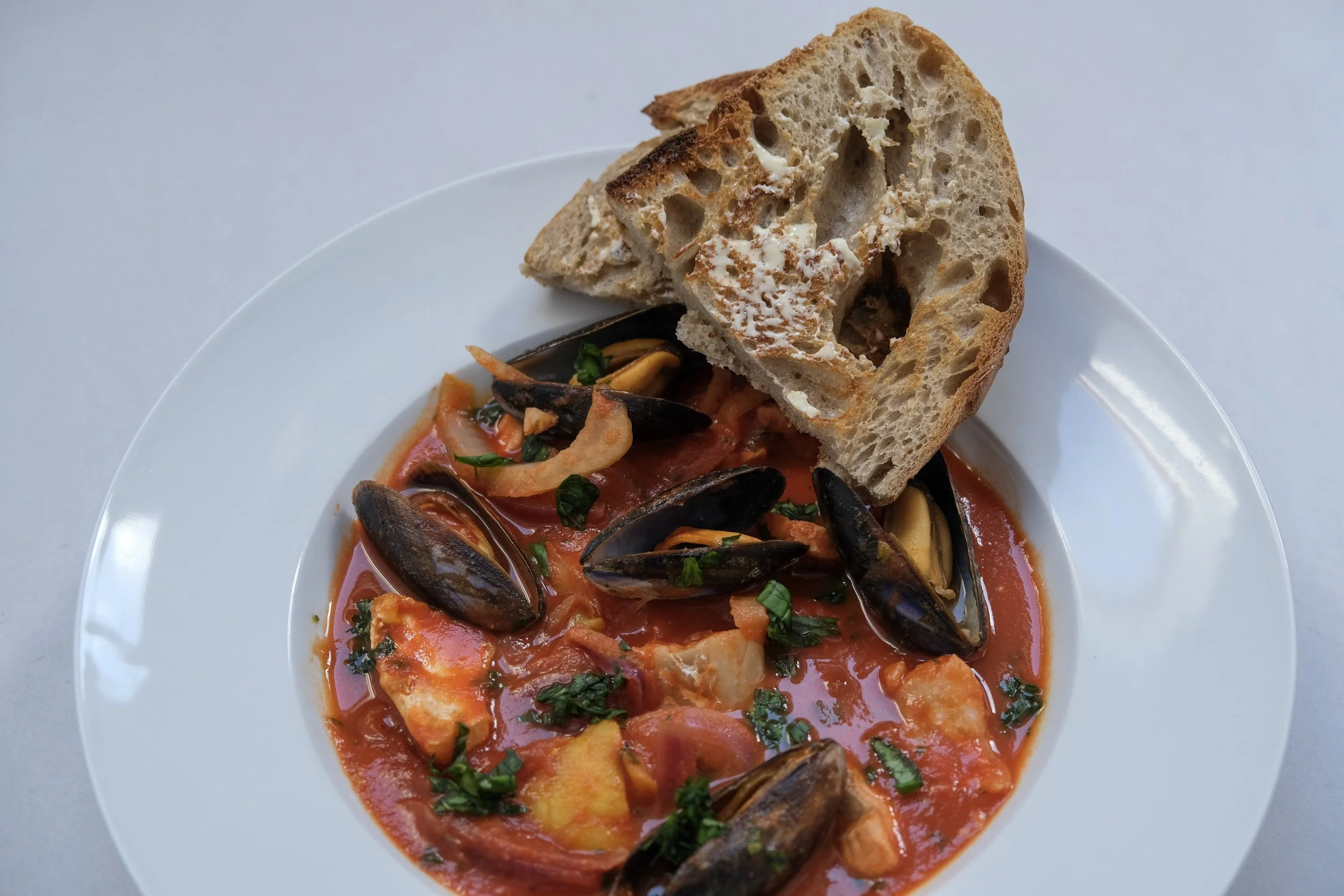 A plated bowl of rich tomato salmon, mussel, and fennel stew topped with fresh parsley and served with two slices of toasted sourdough bread.
