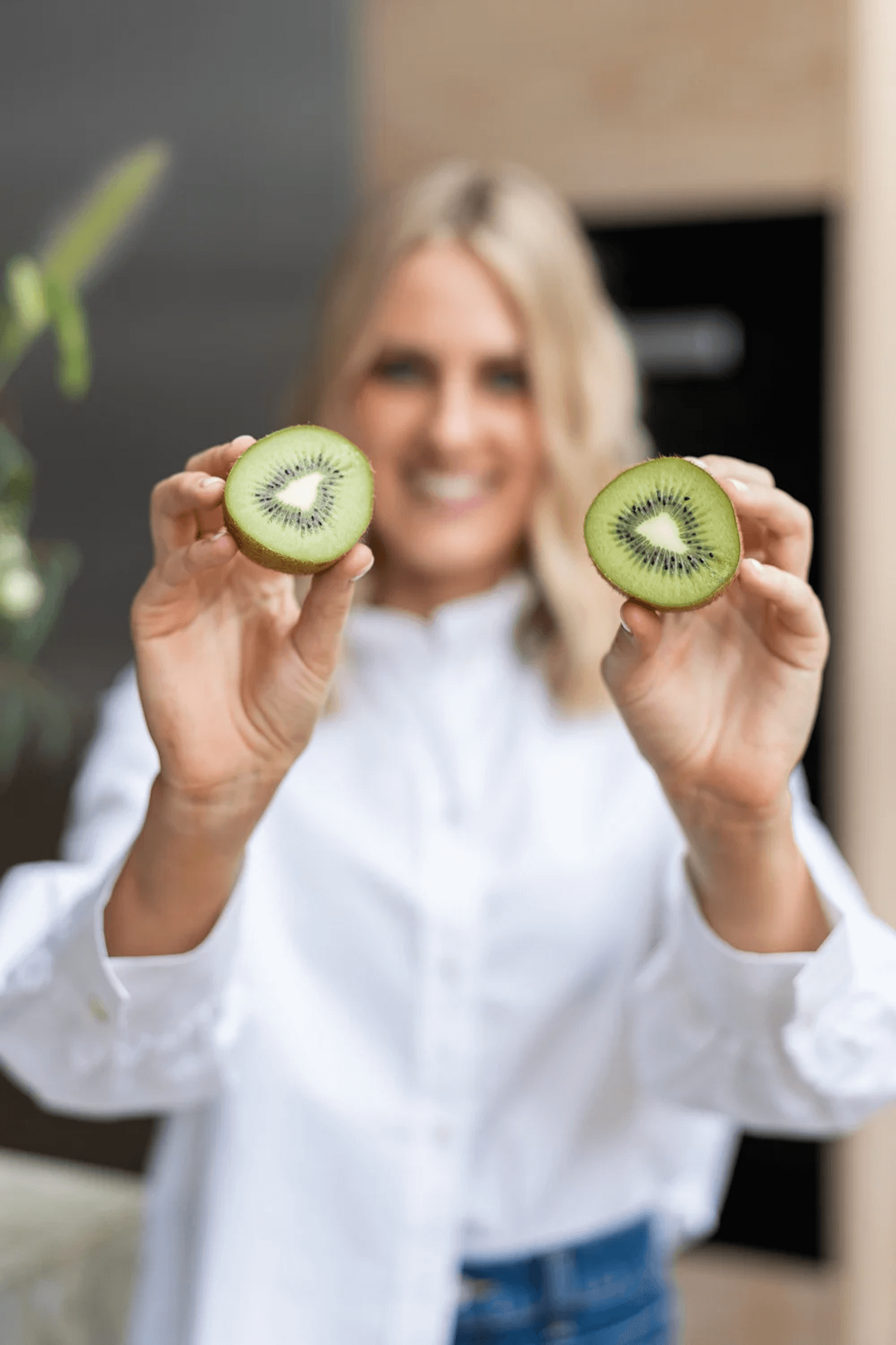A woman smiling and holding two halves of a kiwi fruit, one in each hand.