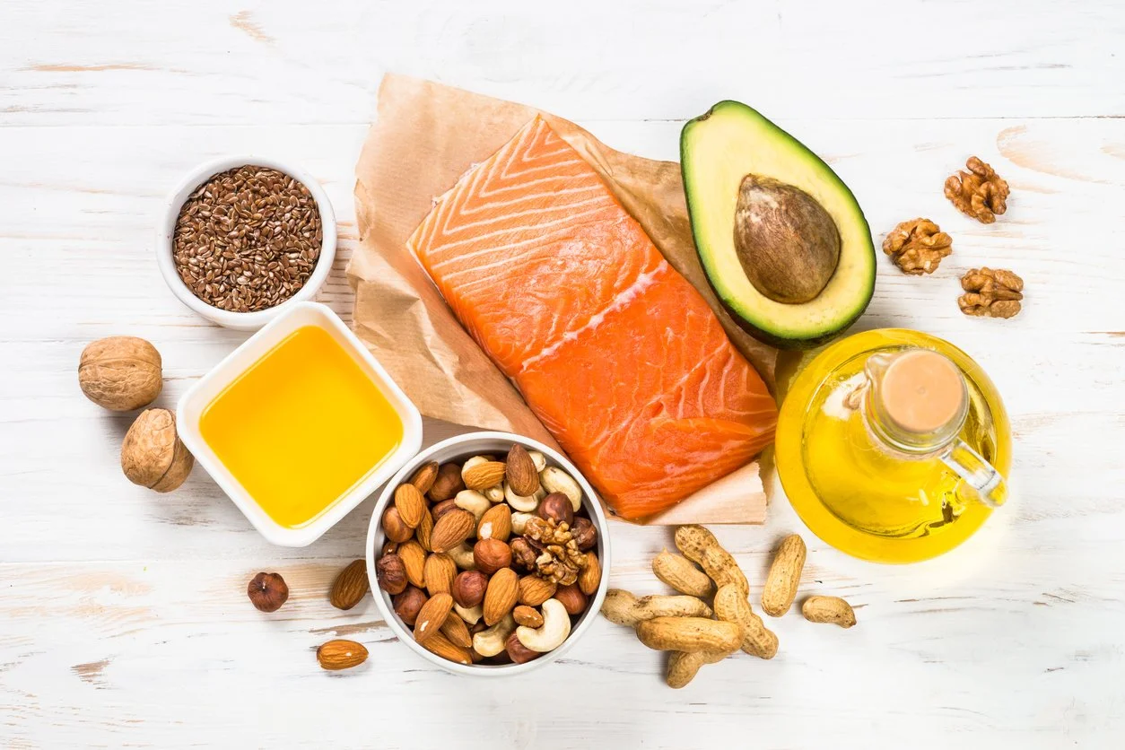 An overhead flat lay of healthy omega 3 rich foods including a salmon fillet, avocado half, olive oil, flaxseeds, and mixed nuts on a white wooden background.