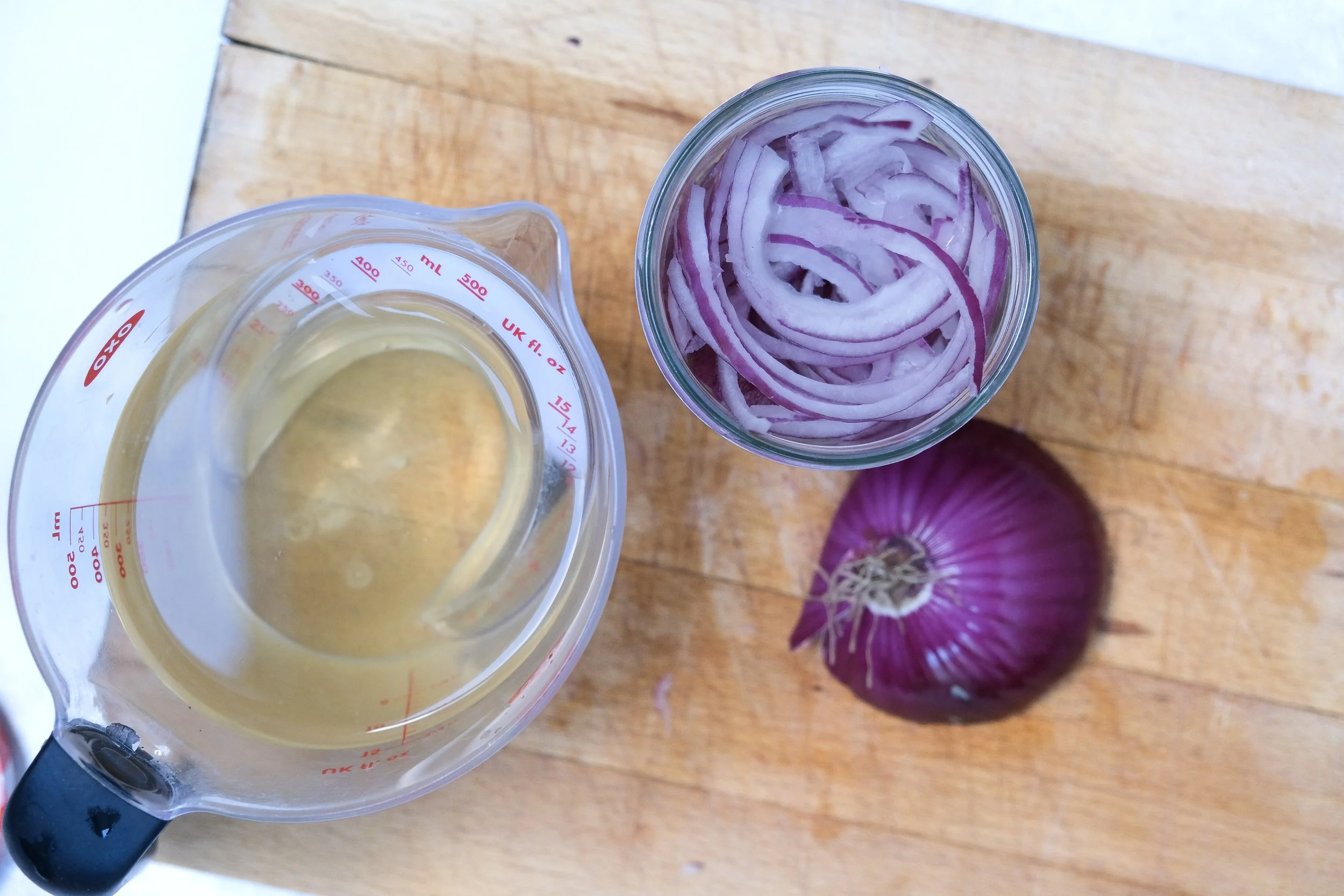 A top-down view of ingredients for pickled red onions, featuring a measuring jug of pickling brine, a glass jar filled with sliced red onions, and half a red onion on a wooden cutting board.