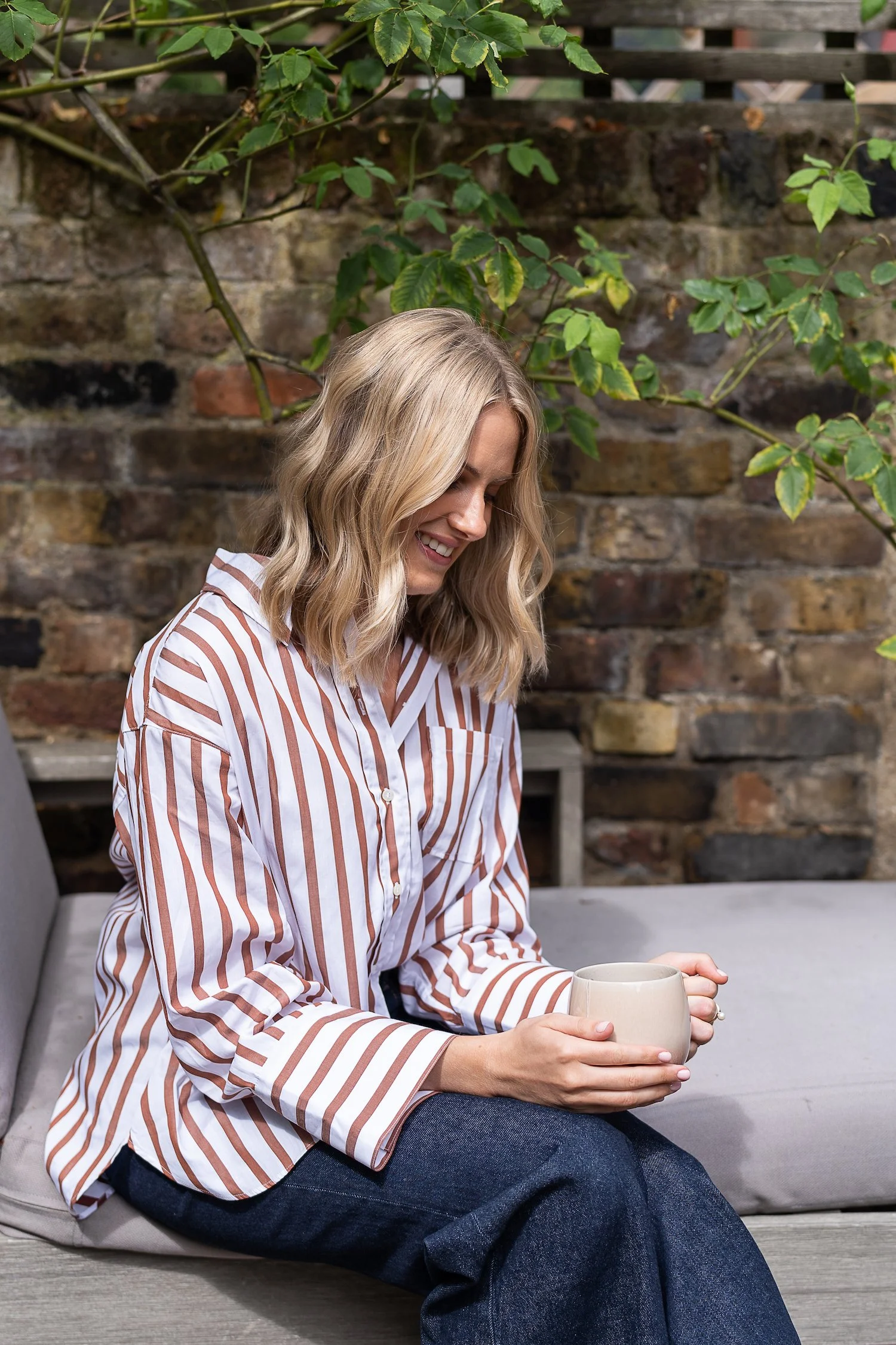 A woman with blonde wavy hair sitting outdoors on a cushioned bench, smiling while holding a beige mug. She is wearing a white and brown striped shirt and dark jeans, with a brick wall and greenery in the background.