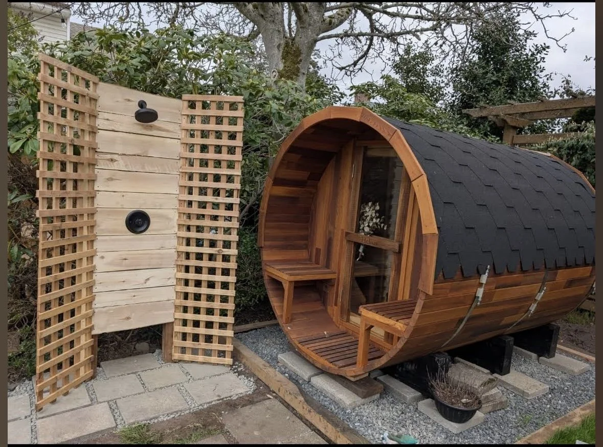 Small wooden barrel-shaped sauna with a curved roof with black shingles, a glass door, and a small bench outside. To the left, a wooden outdoor shower with a showerhead and a speaker, set on a paved stone patio surrounded by greenery.