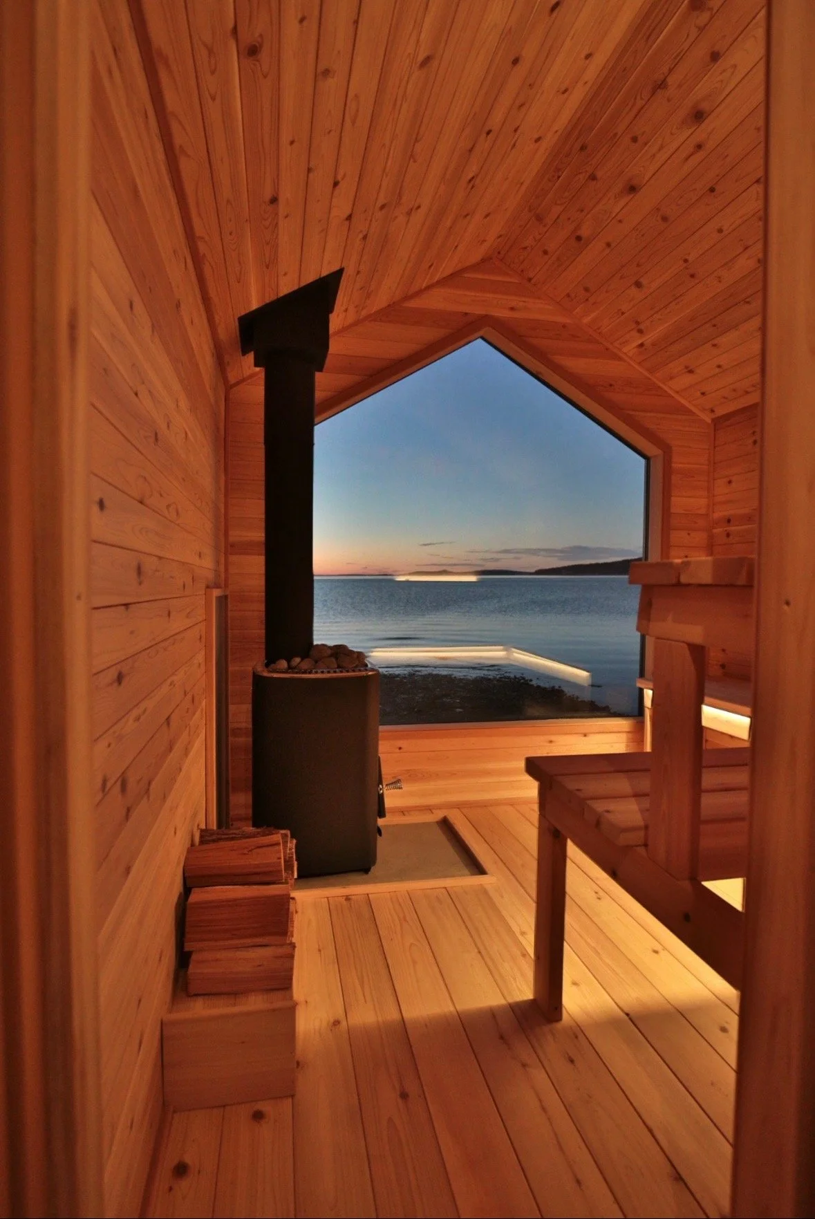 Interior view of a wooden sauna with a view of a body of water and a sunset through a large window.