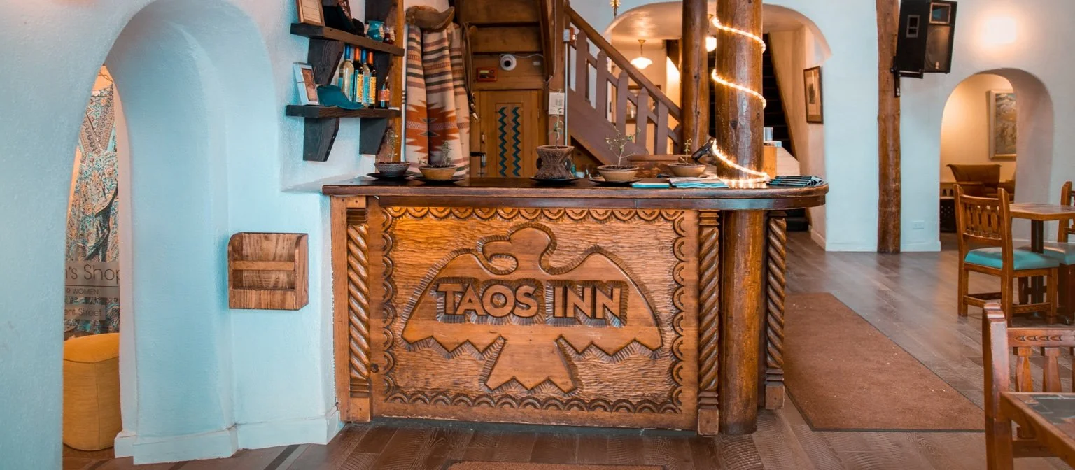 Interior of TAOS INN with carved wooden front desk and wooden staircase, books on shelves, and cozy decor.