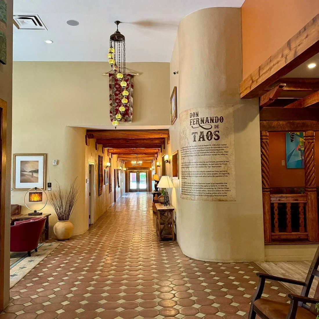 Hotel interior hallway with tiled floor, artwork on walls, and wooden accents. There are lamps and a large decorative vase with dry plants near a red armchair, leading to a glass double door at the end of the hall.