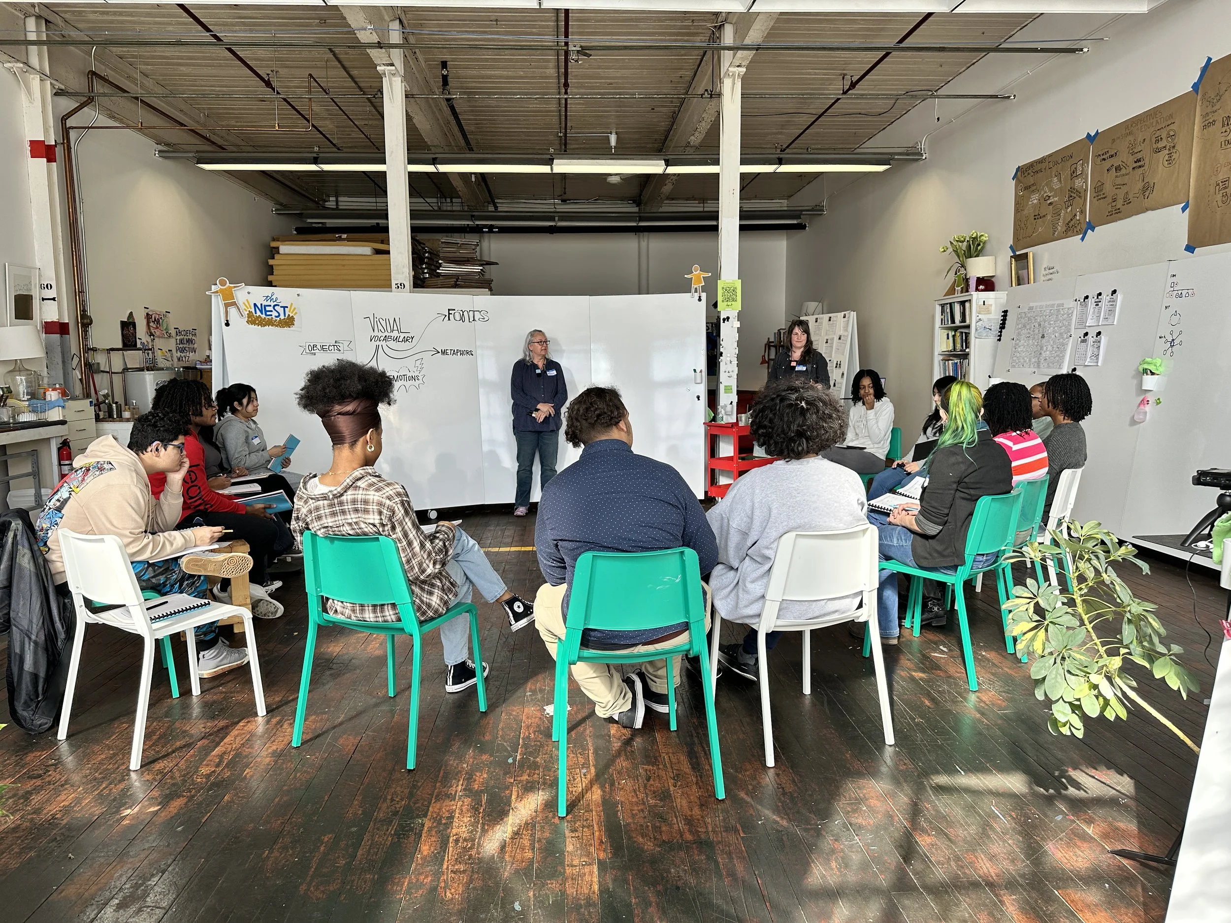 A diverse group of students attending a classroom lecture or workshop in a bright, industrial-style room with whiteboards and bookshelves. Two instructors are standing at the front, while the students sit in colorful chairs, listening and taking notes.