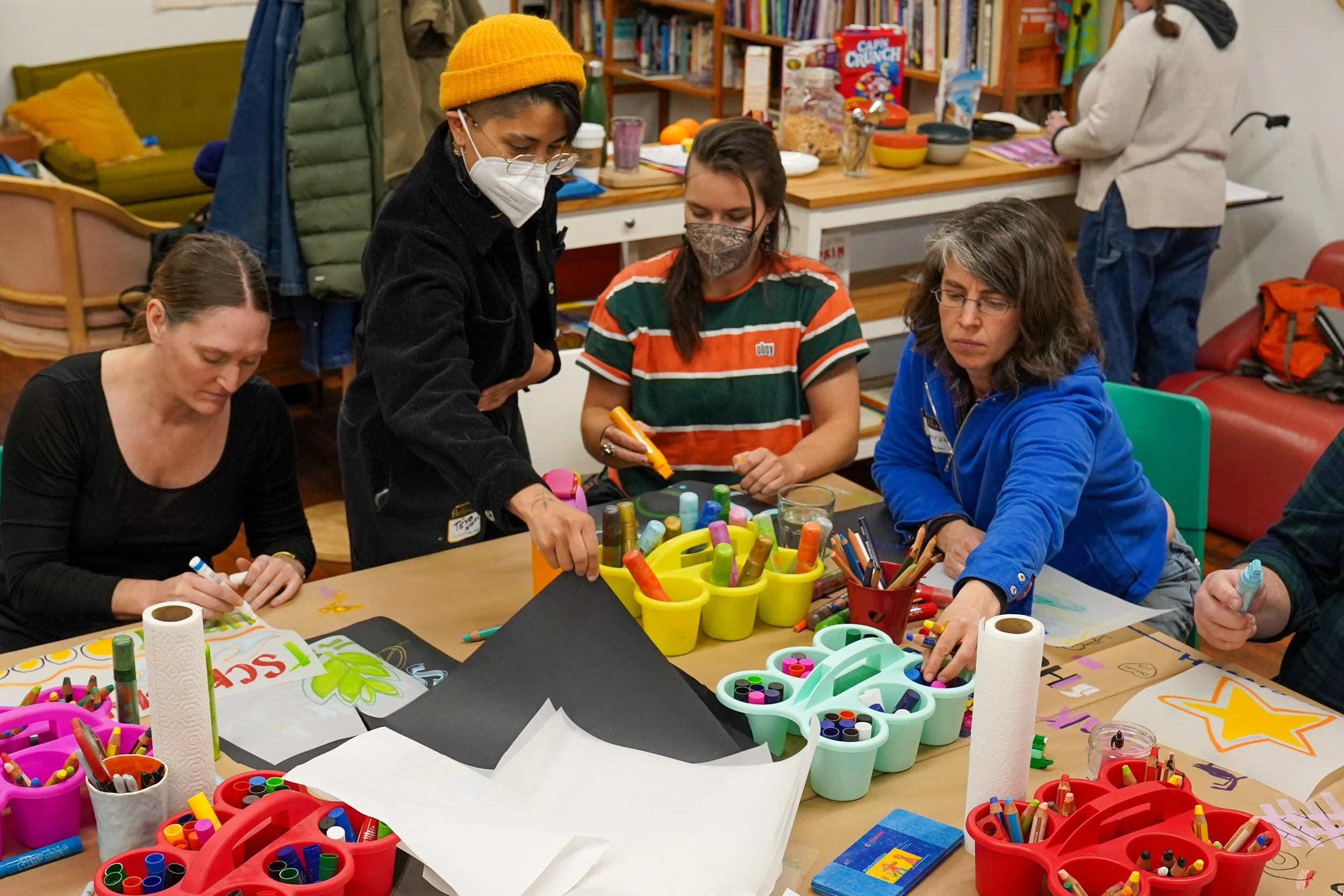 Group of people gathered around a table engaging in arts and crafts activities, surrounded by colorful markers, paper, and art supplies.
