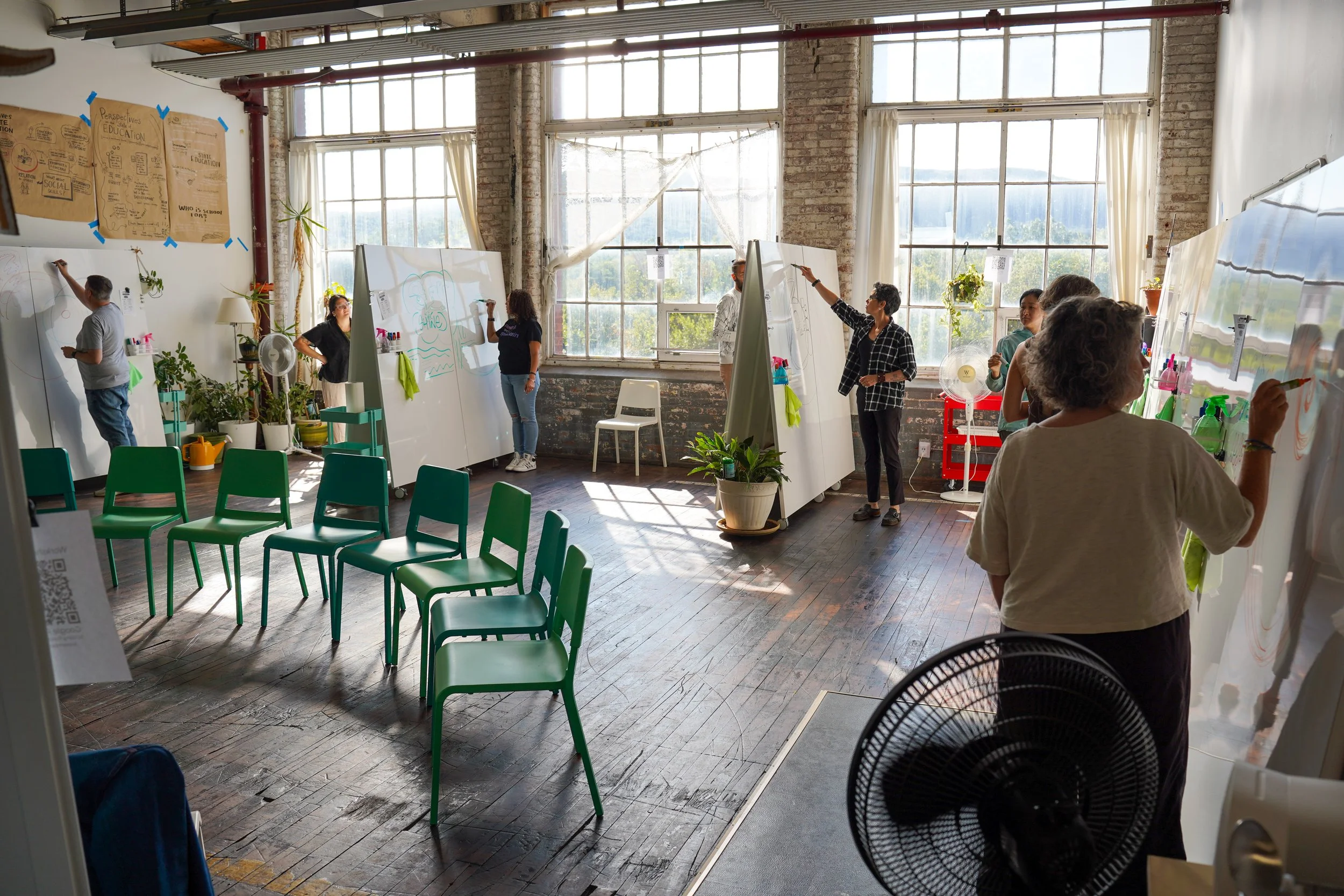 People participating in a whiteboard drawing activity in an art classroom with large windows, potted plants, and green chairs arranged in rows.
