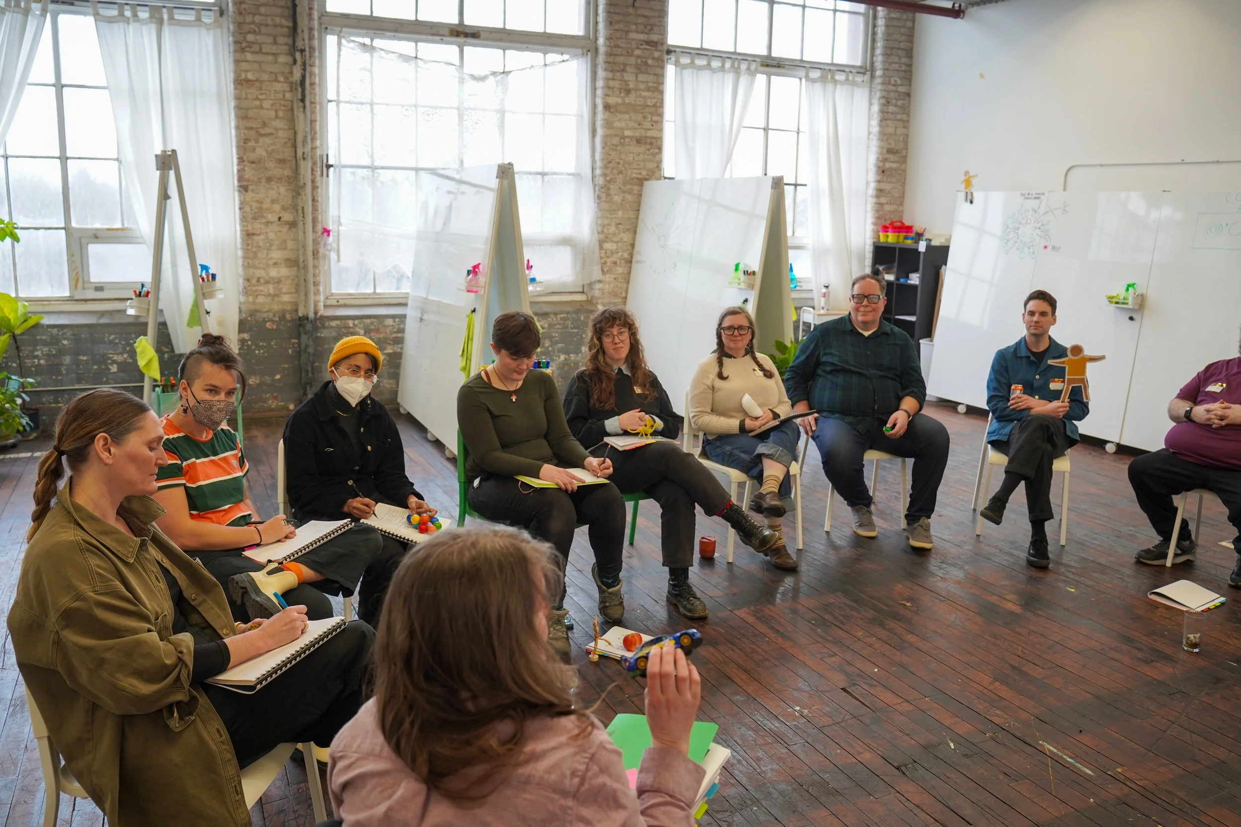 A group of people is seated in a semi-circle in a bright, spacious room with large windows, participating in a discussion or workshop. Some are holding notebooks, pens, or small models, and many wear masks.