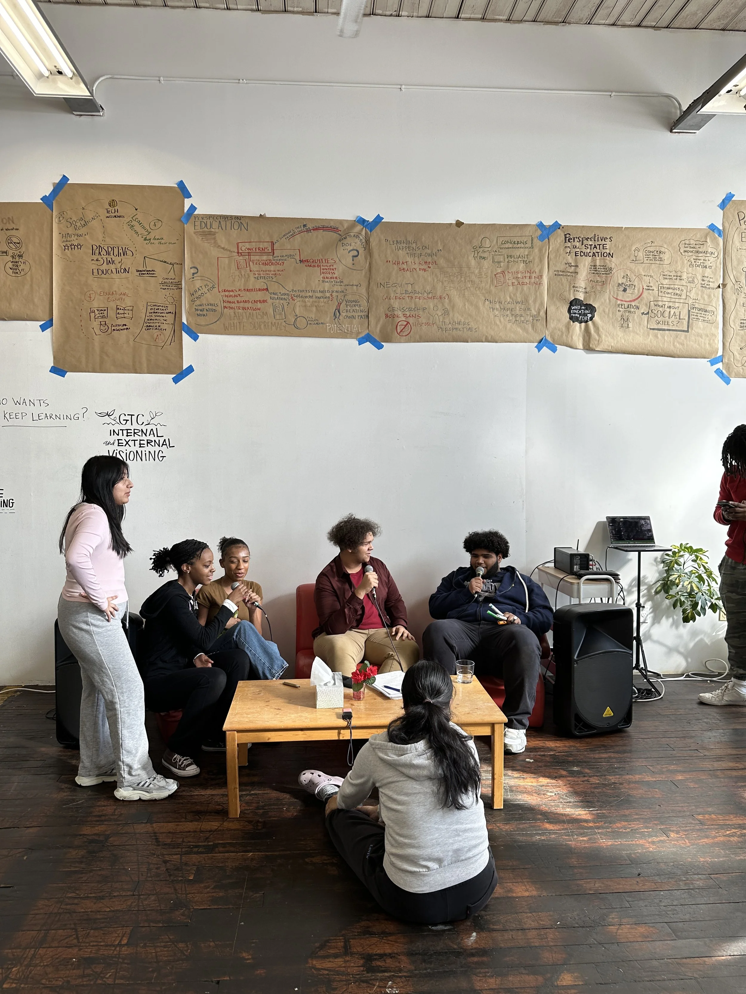 Group of five young people participating in a panel discussion, with three speaking into microphones while standing or sitting on a red sofa, and two others listening, one sitting on the floor with back to the camera and the other standing on the right side. Behind them is a white wall with large brown paper sheets with handwritten notes and diagrams taped to it.