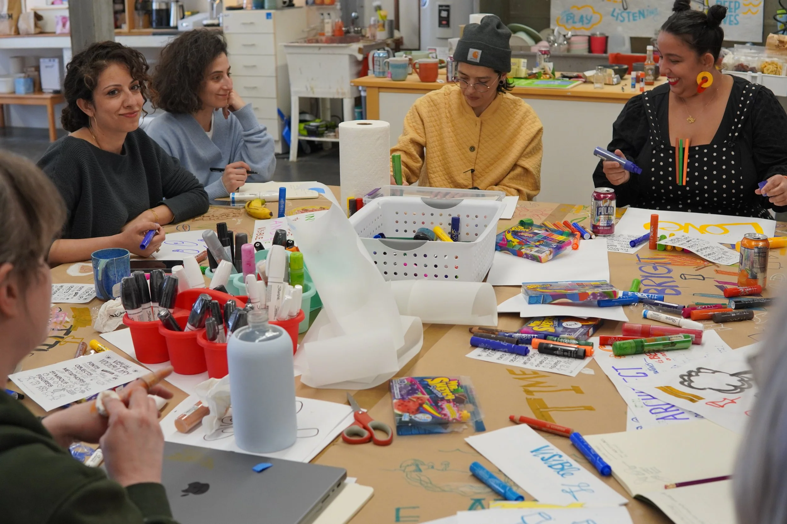 A group of people sitting around a table filled with markers, paper, and art supplies, engaging in a creative activity in a classroom or workshop setting.