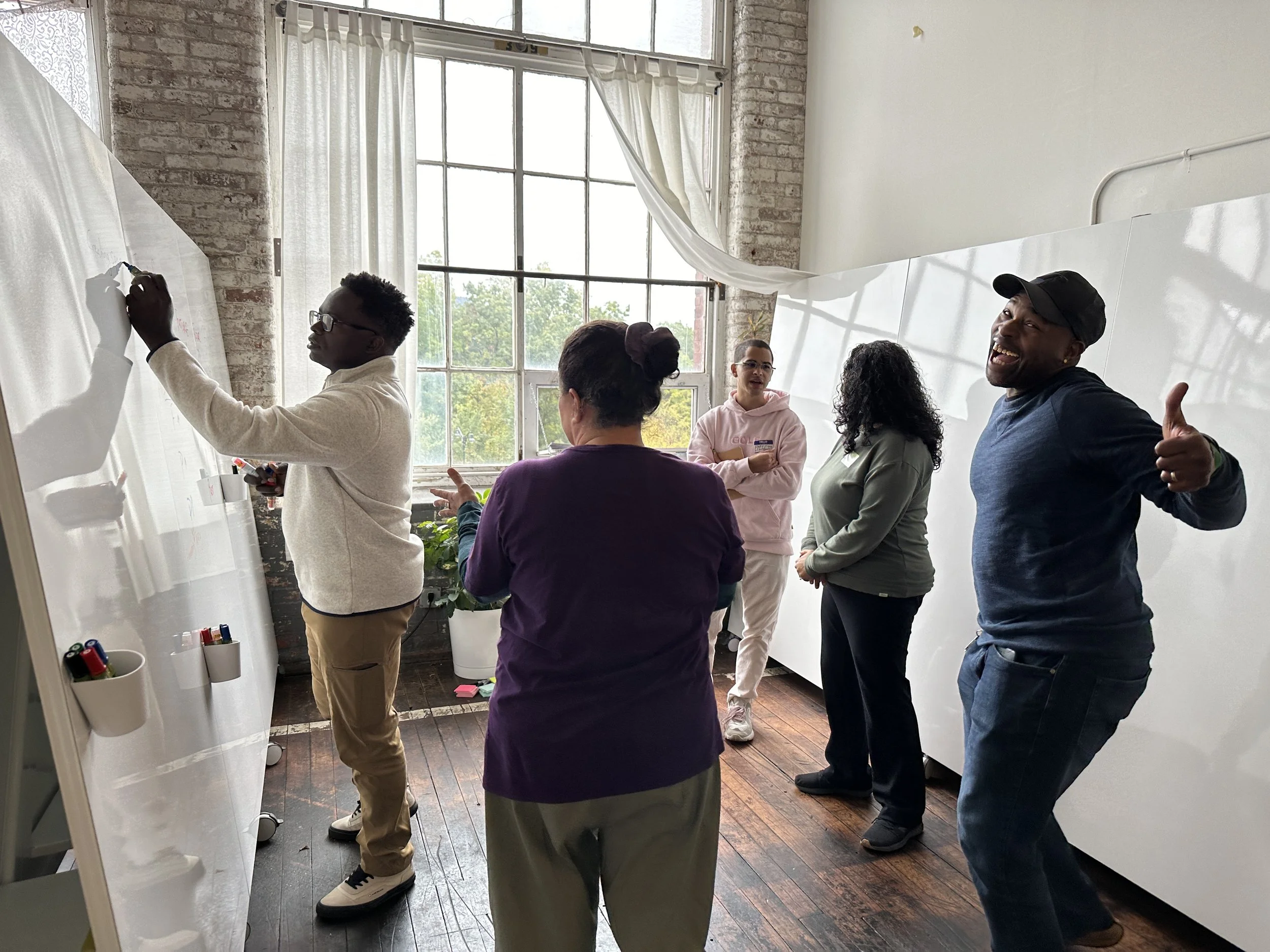 Group of five people engaged in a discussion or workshop in a bright room with large windows and whiteboards.