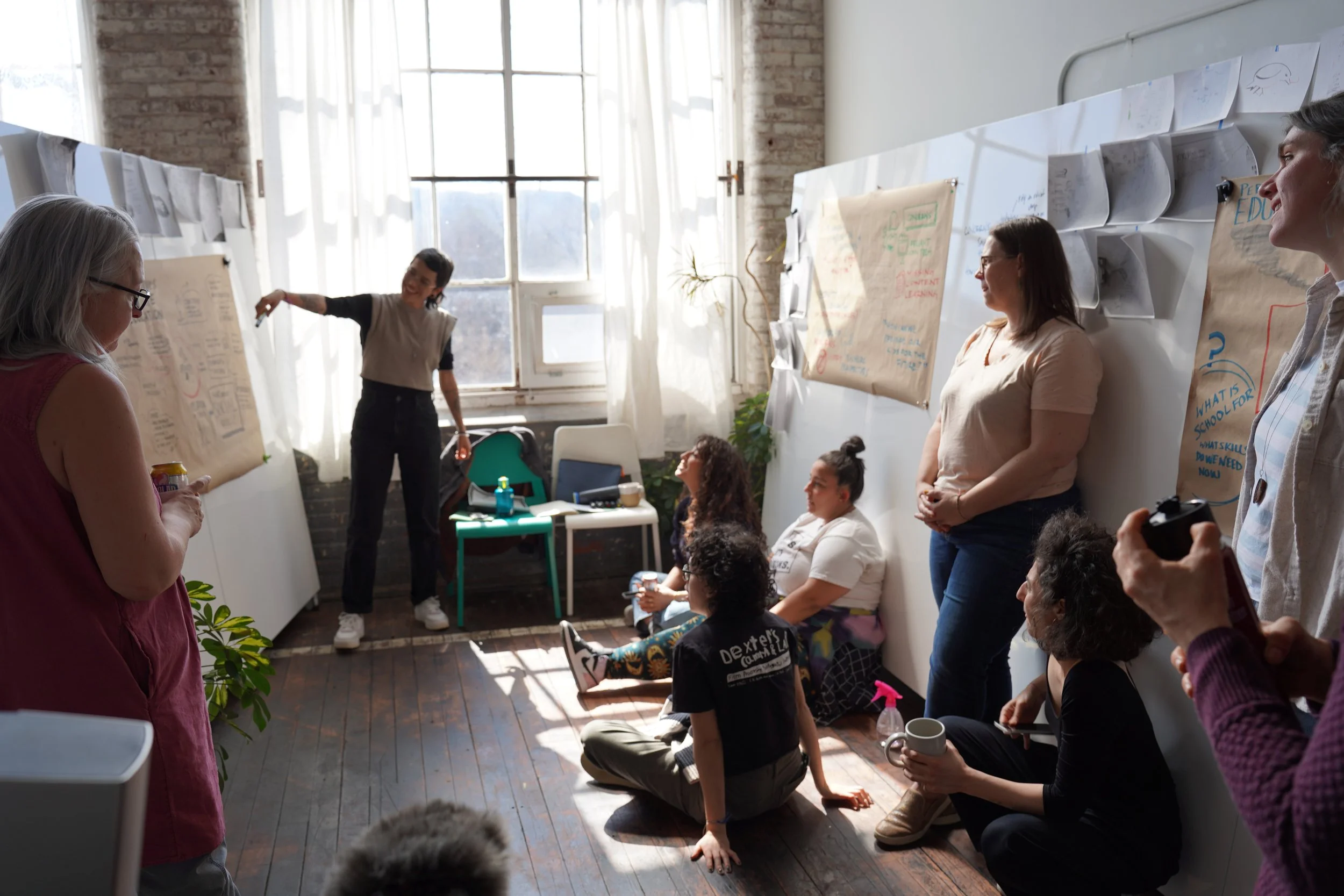Group of people in a workshop room with large papers and whiteboard, one person is presenting while others sit and stand, sunlight coming through large window.