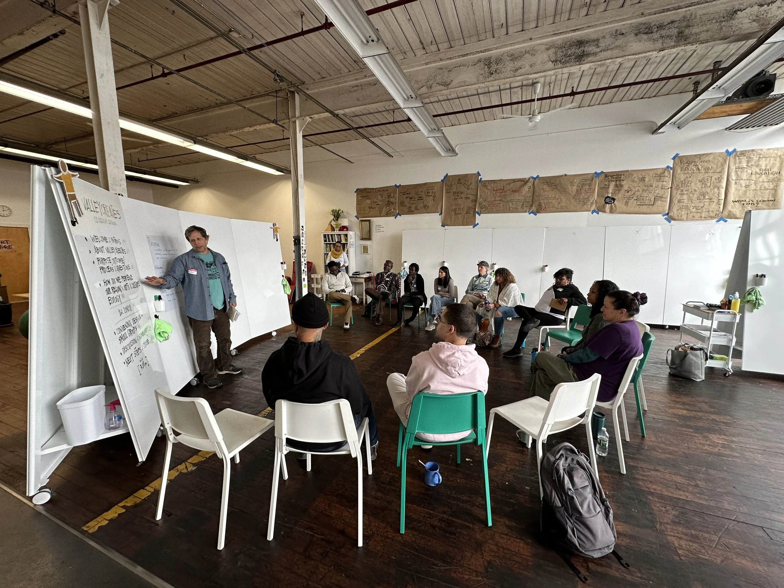 A group of people participating in a workshop or meeting. A person is standing near a large whiteboard at the front, addressing the group. The participants are seated around in a semi-circle, listening attentively. The room has high ceilings with exposed pipes and a white wall with large paper sheets taped up. The floor is wooden, and there is a cart with supplies on the right side.