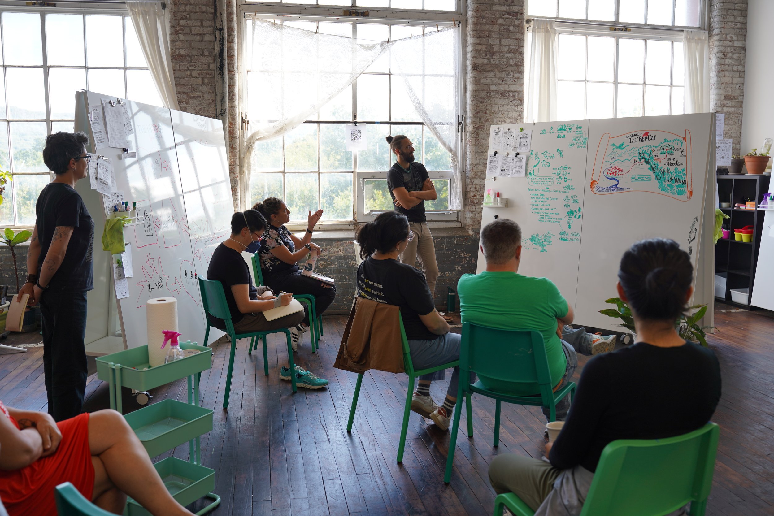 A group of people attending a workshop or class in a bright room with large windows and exposed brick walls. Some are seated, taking notes or listening, while an instructor stands near large whiteboards filled with illustrations and notes.