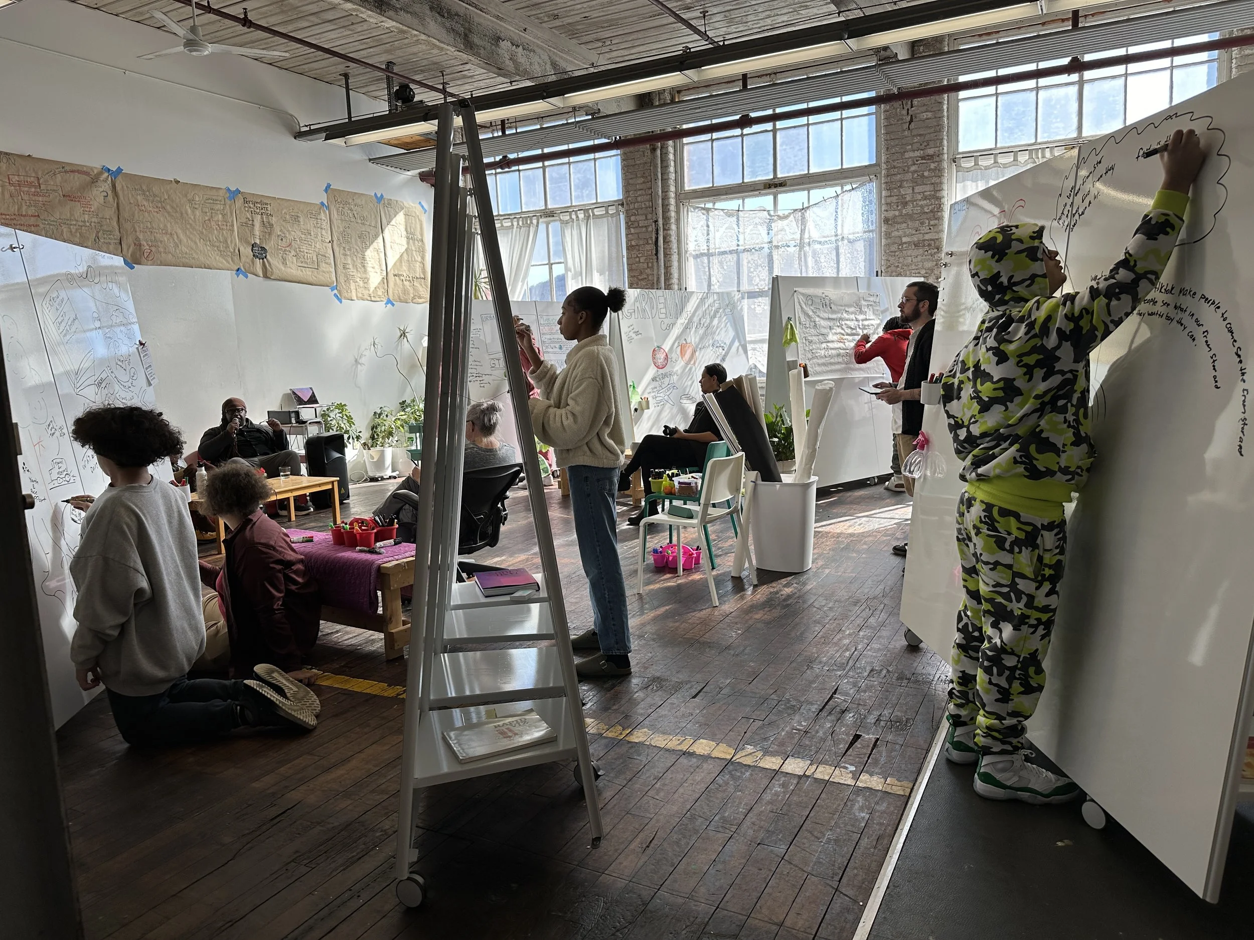 People engaged in a collaborative workshop or brainstorming session in a bright, industrial-style studio with large windows, whiteboards, and assorted materials.