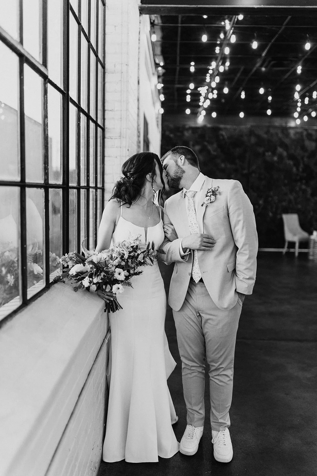 A black and white photo of a bride and groom sharing a kiss indoors. The bride is holding a bouquet of flowers and wearing a sleeveless dress, while the groom is dressed in a light-colored suit with a patterned tie. They are standing near a large window with a grid pattern, with string lights hanging from the ceiling in the background.