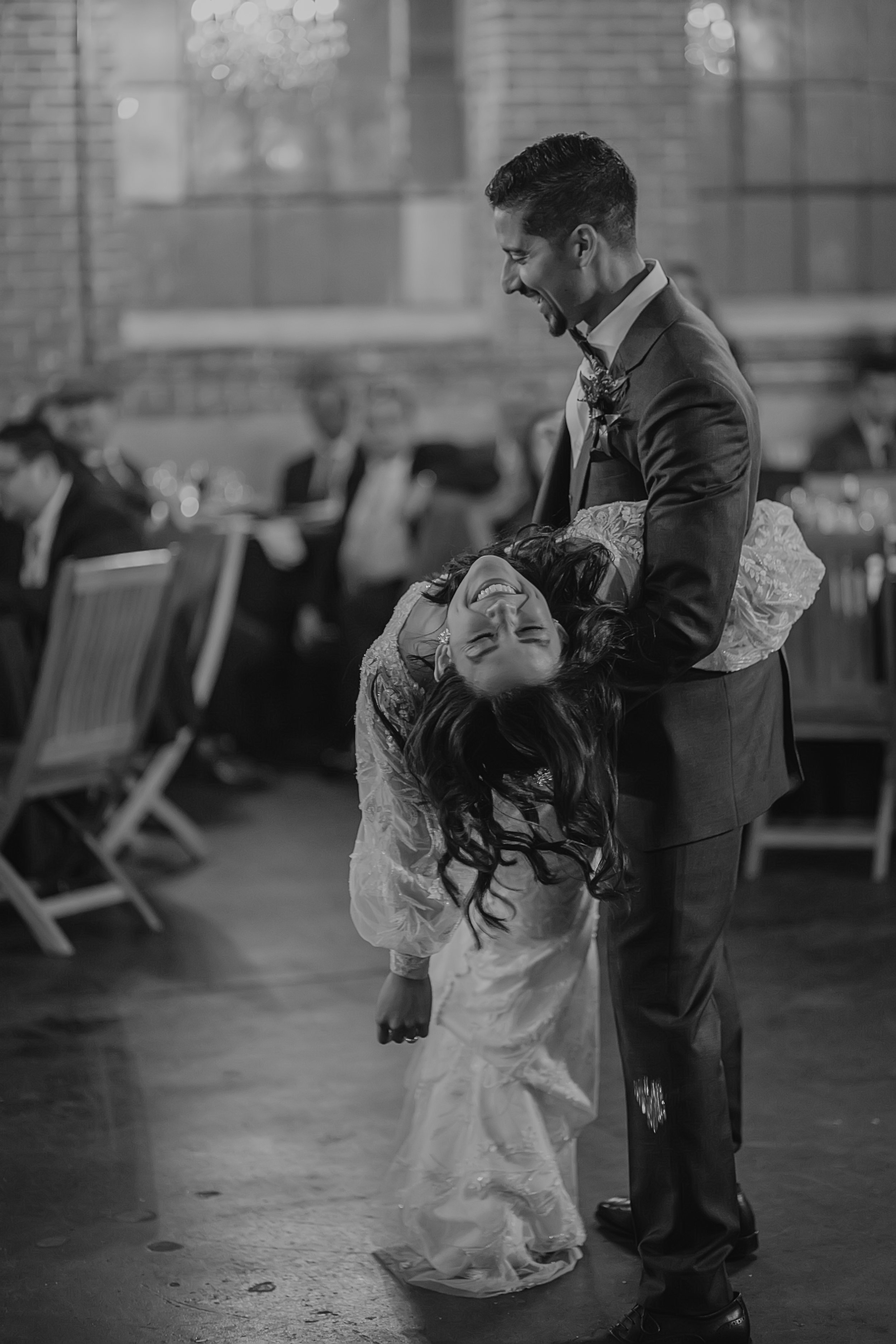 Moss Denver A man in a tuxedo holding a woman in a wedding dress, she is bent backwards with her eyes closed and a big smile, at a wedding reception with guests seated at tables in the background.