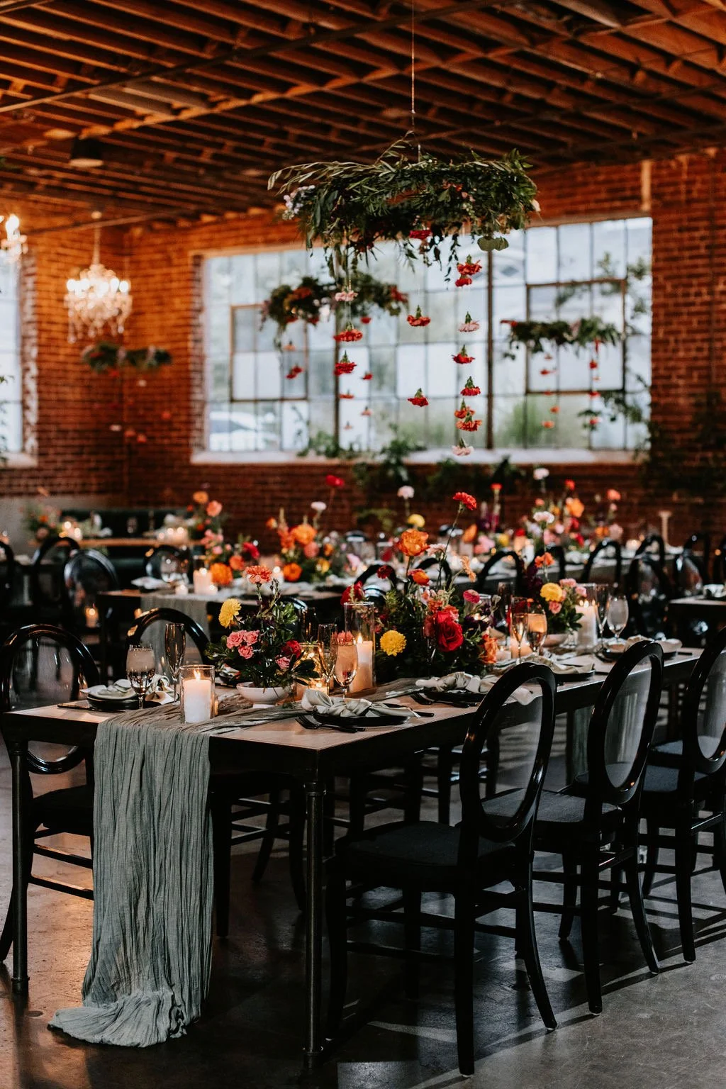 Elegant indoor wedding reception setup at Moss Denver with long tables adorned with colorful floral centerpieces, candles, and glassware, in a rustic brick-walled venue with large windows and hanging floral arrangements. Wedding planner Denver CO