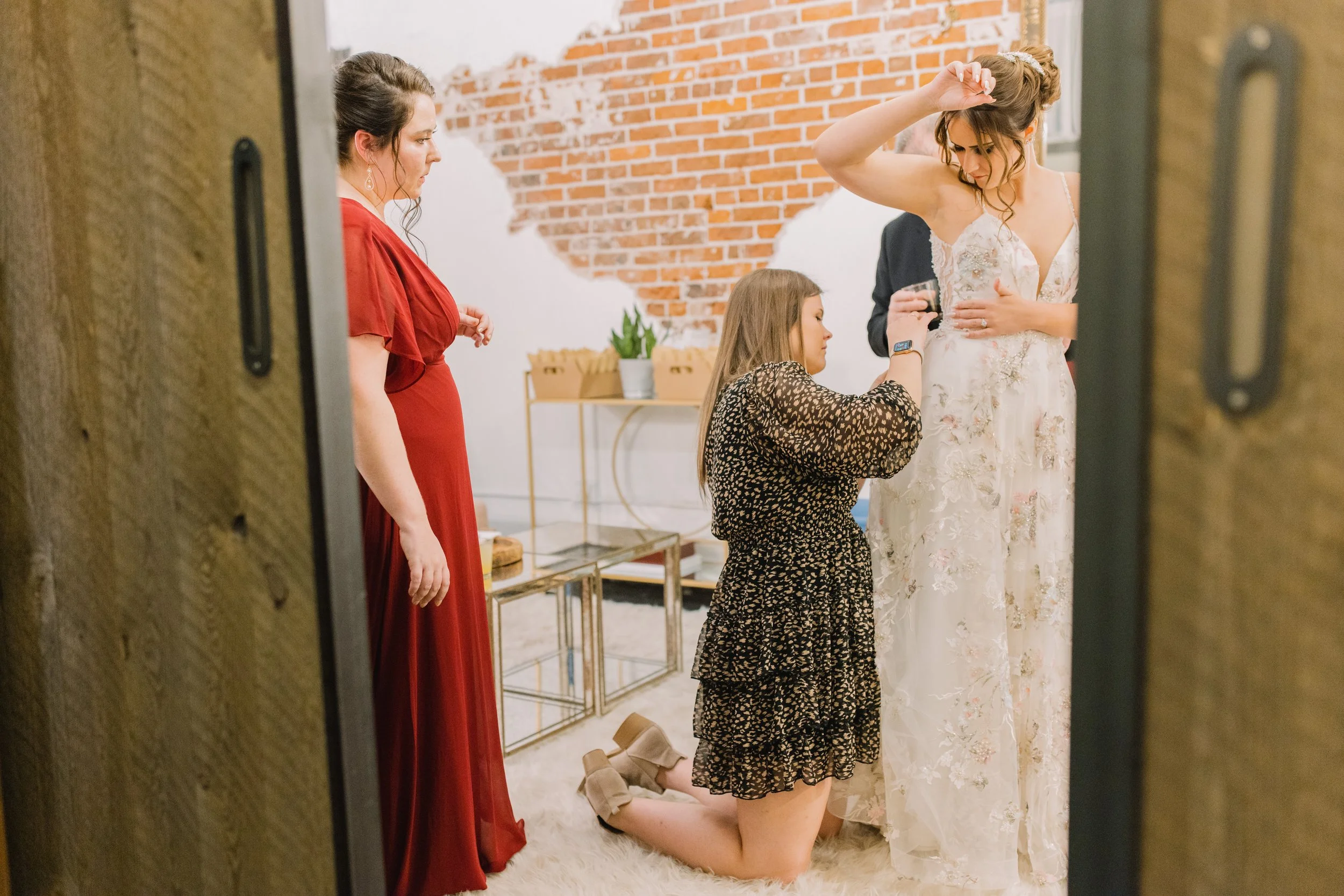 A woman in a white wedding dress is getting ready, assisted by a woman kneeling and applying makeup or powder to her face, while another woman in a red dress observes, all inside a room with an exposed brick wall.