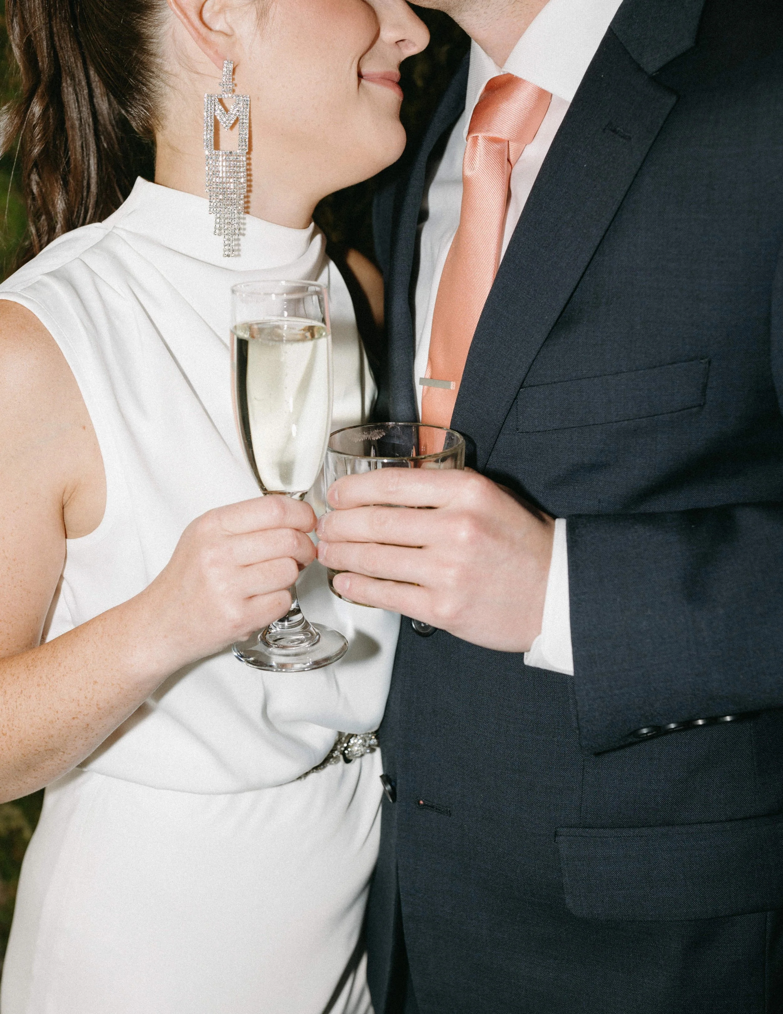Moss Denver Coordinator Close-up of a couple at a celebration, holding glasses of champagne and wine, dressed in formal attire, with the woman leaning in close to the man.