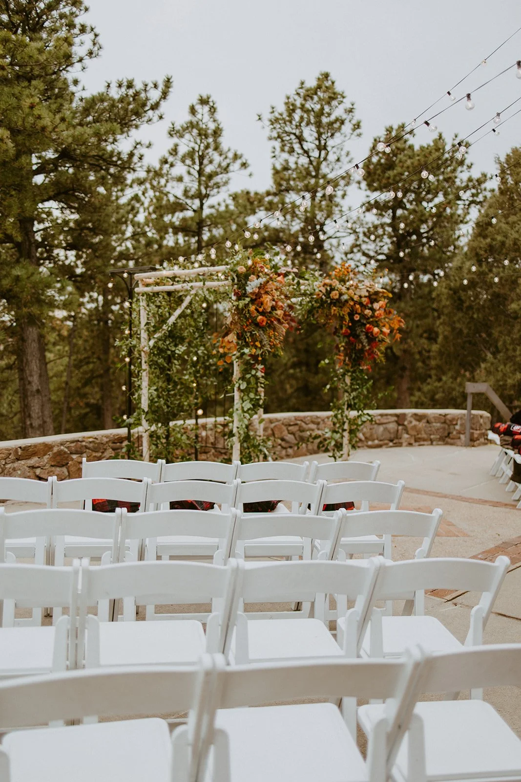 Outdoor wedding ceremony setup at Boettcher Mansion Golden, Colorado with white chairs and floral arch in a wooded area, decorated with string lights. Professional wedding planner Colorado Springs Golden
