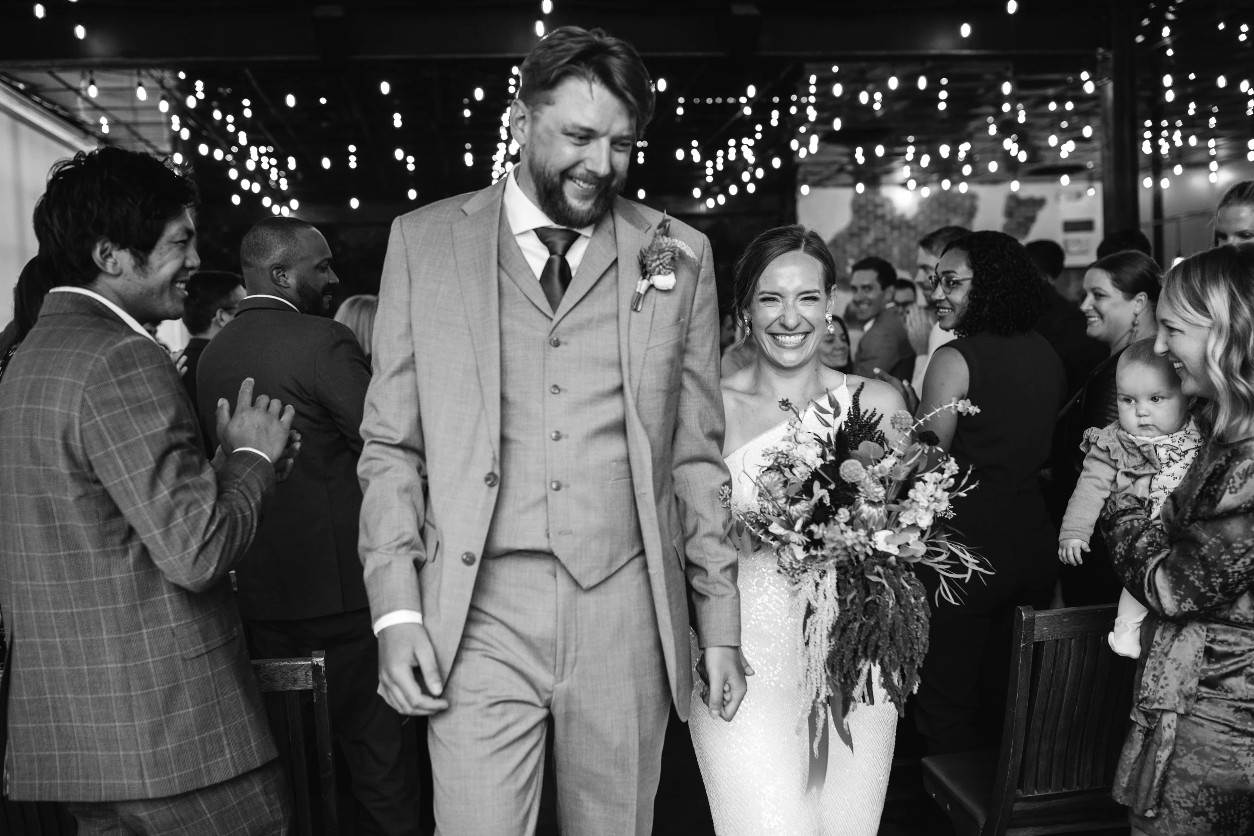 Black and white photo of a wedding reception with smiling bride and groom holding hands, surrounded by guests, some clapping and others taking photos, with string lights overhead.