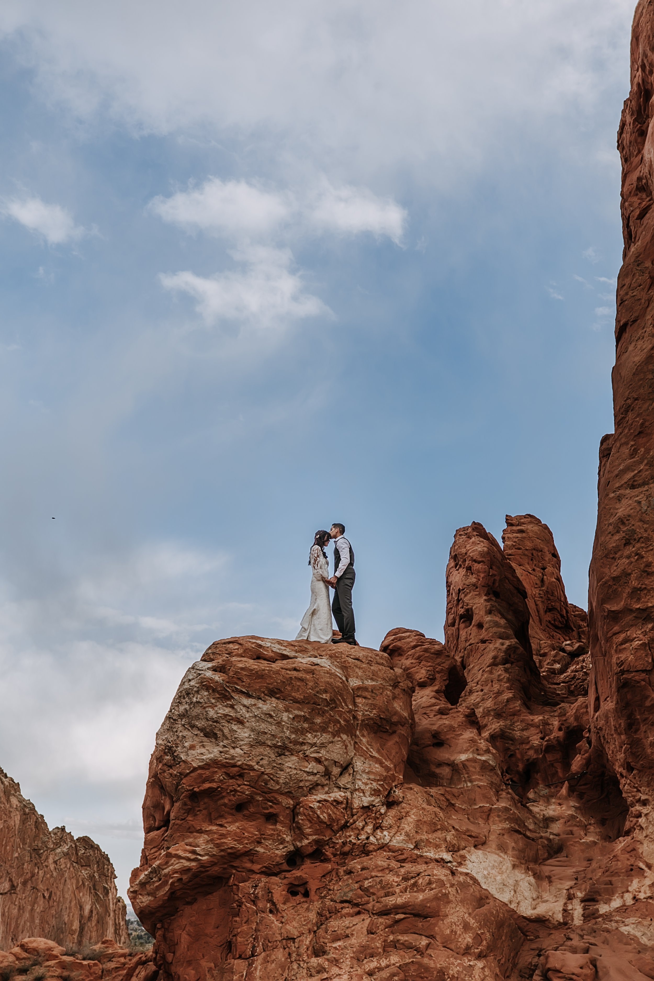 Garden of the God's Wedding Planner A couple dressed in wedding attire standing on a large rock outcrop among red rock formations, kissing under a cloudy sky.