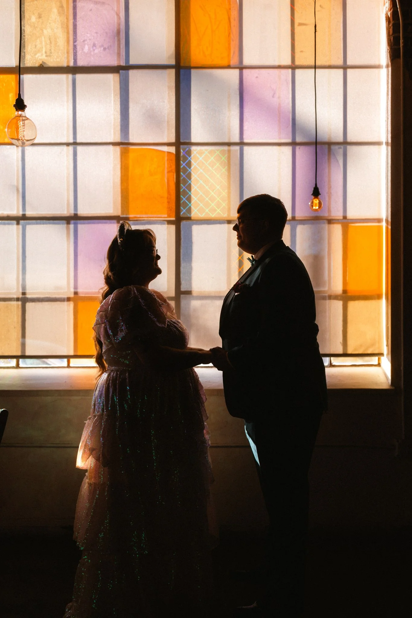 Silhouette of a couple holding hands at Moss Denver Wedding Planner, standing in front of a colorful stained glass window, possibly during a wedding or special event.