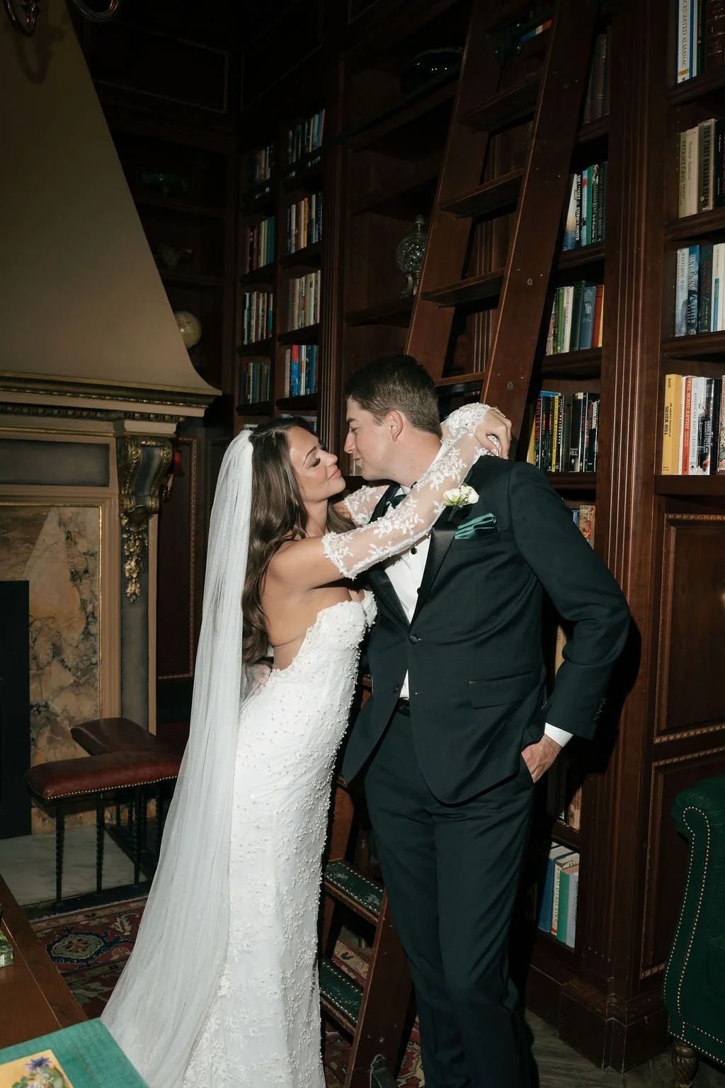 A bride and groom at The Broadmoor Hotel Colorado Springs Wedding Planner in a warmly lit room with dark wood bookshelves filled with books, a fireplace, and a collage of decorative objects.