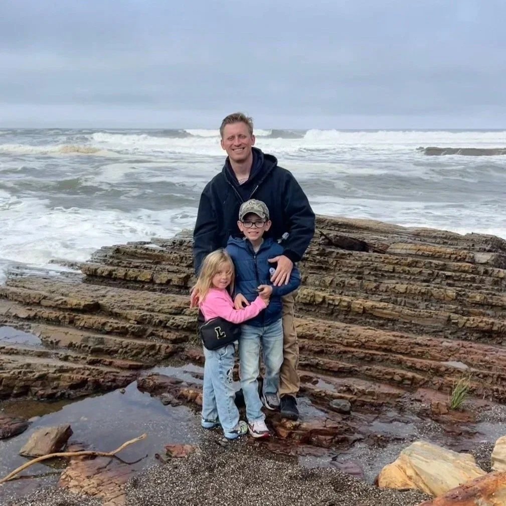 A family of three standing on rocks at the beach with ocean waves in the background, on a cloudy day.