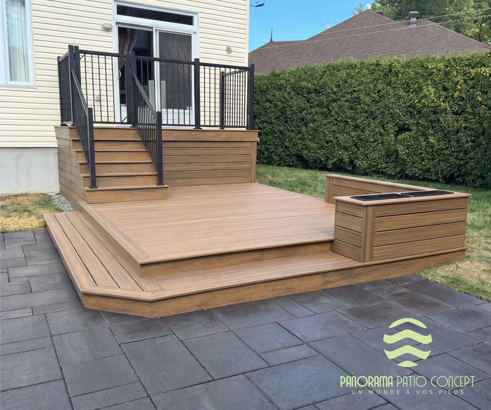  Terrasse en bois avec escalier menant à une porte-fenêtre, entourée d'une maison avec une façade beige, un jardin et un mur de haie verte, logo de Panorama Patio Concept en bas à droite.