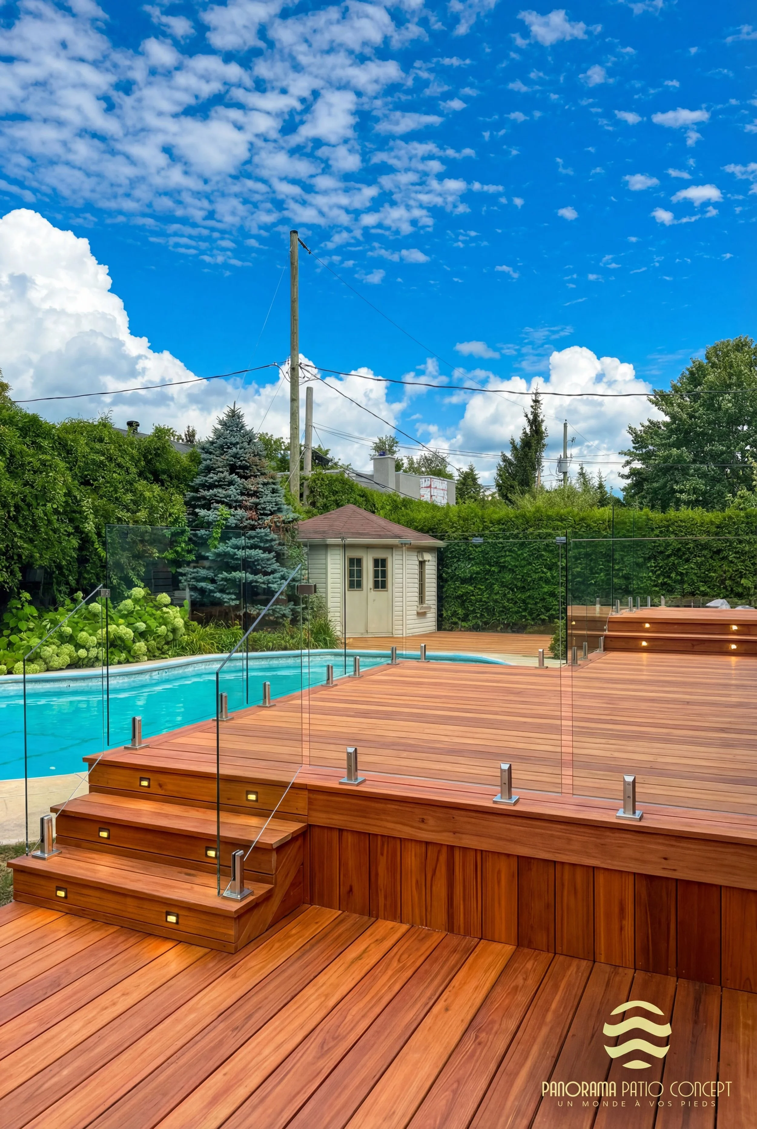 Terrasse en bois avec escalier lumineux, piscine et vue sur jardin sous ciel bleu avec nuages.