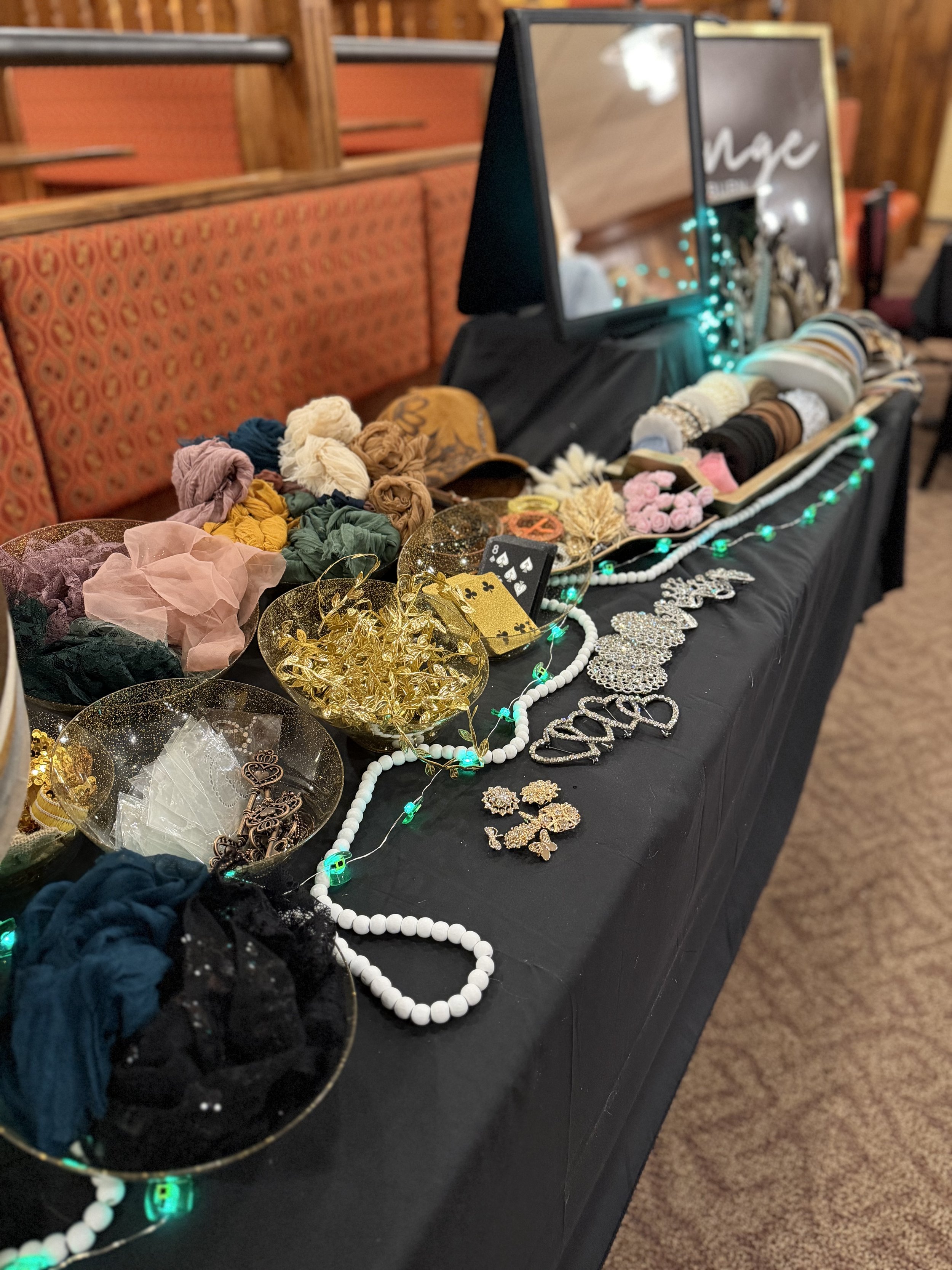 A display table with various jewelry items, headbands, scrunchies, and decorative accessories, some in bowls, with a mirror and a sign in the background, set in a warm-toned room with wooden paneling.