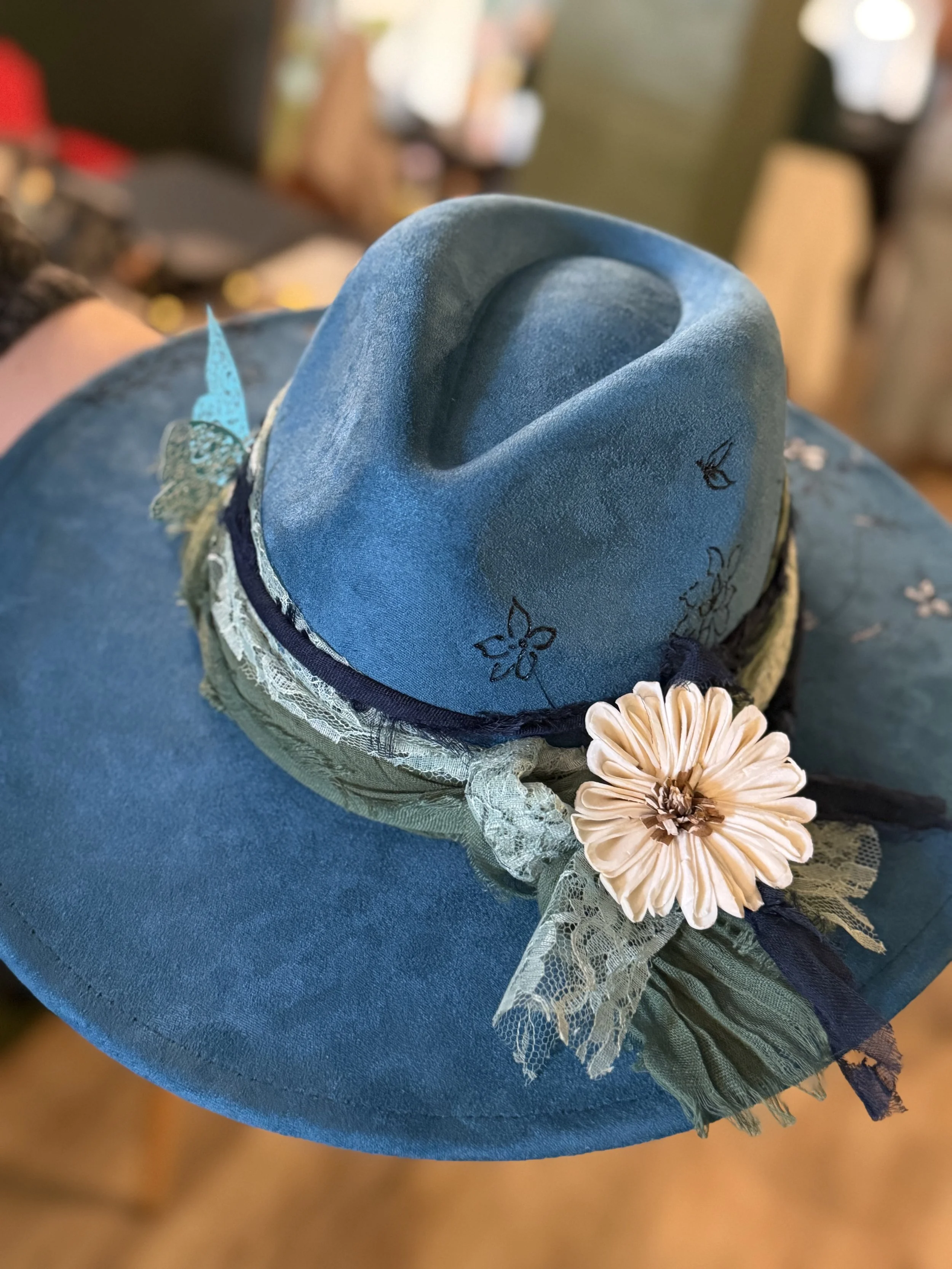 A close-up of a decorative blue hat with embroidered floral details, lace trimmings, and a cream-colored fabric flower, resting on a blue surface with a blurred background.