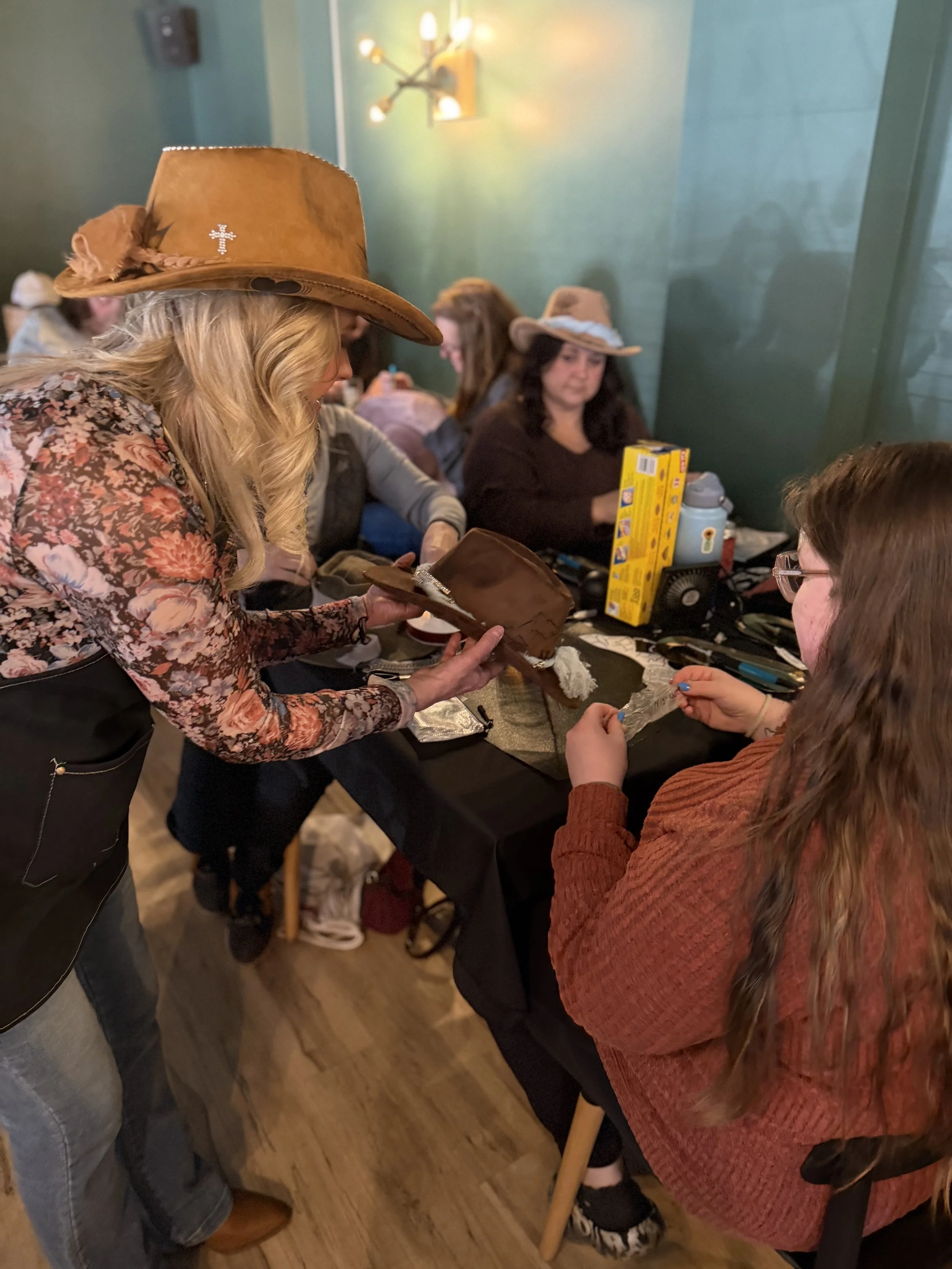 A group of women wearing cowboy hats sitting and standing around a table at a gathering or party, with one woman serving food or a dessert.
