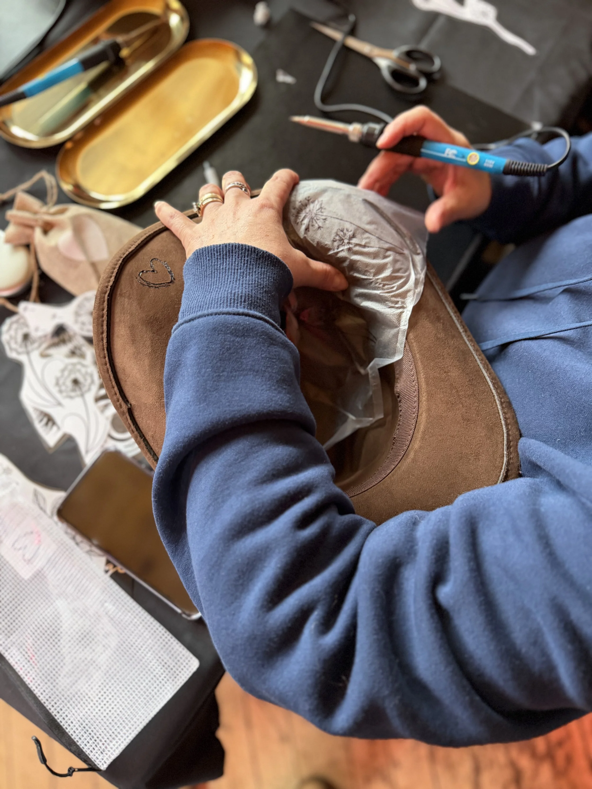 Person holding a heated soldering iron in one hand and working on a piece of fabric or paper with the other hand, with various tools and materials on a black table.
