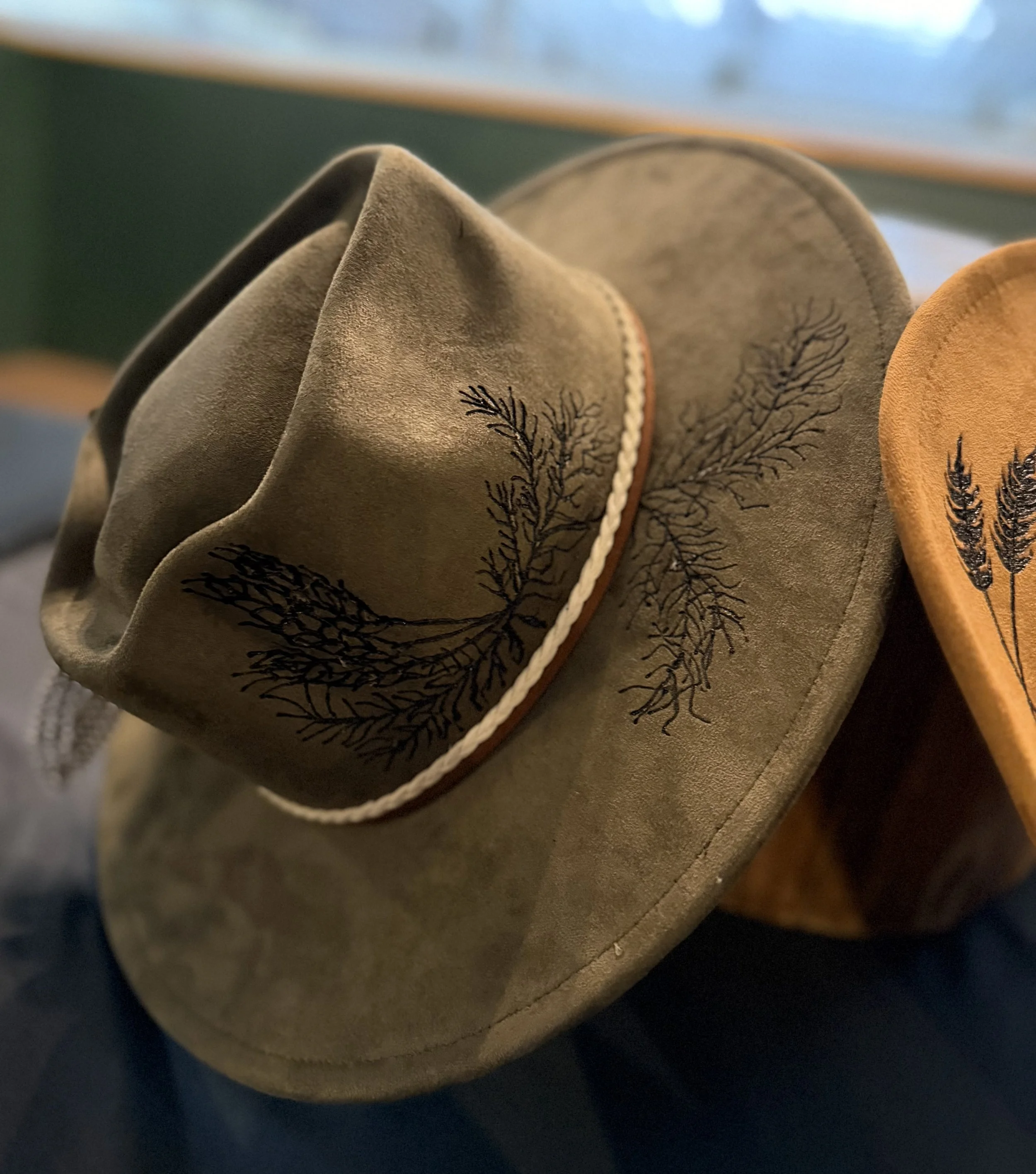 Close-up image of three suede cowboy hats with embroidered black trees, stacked on a surface with a blurred background.