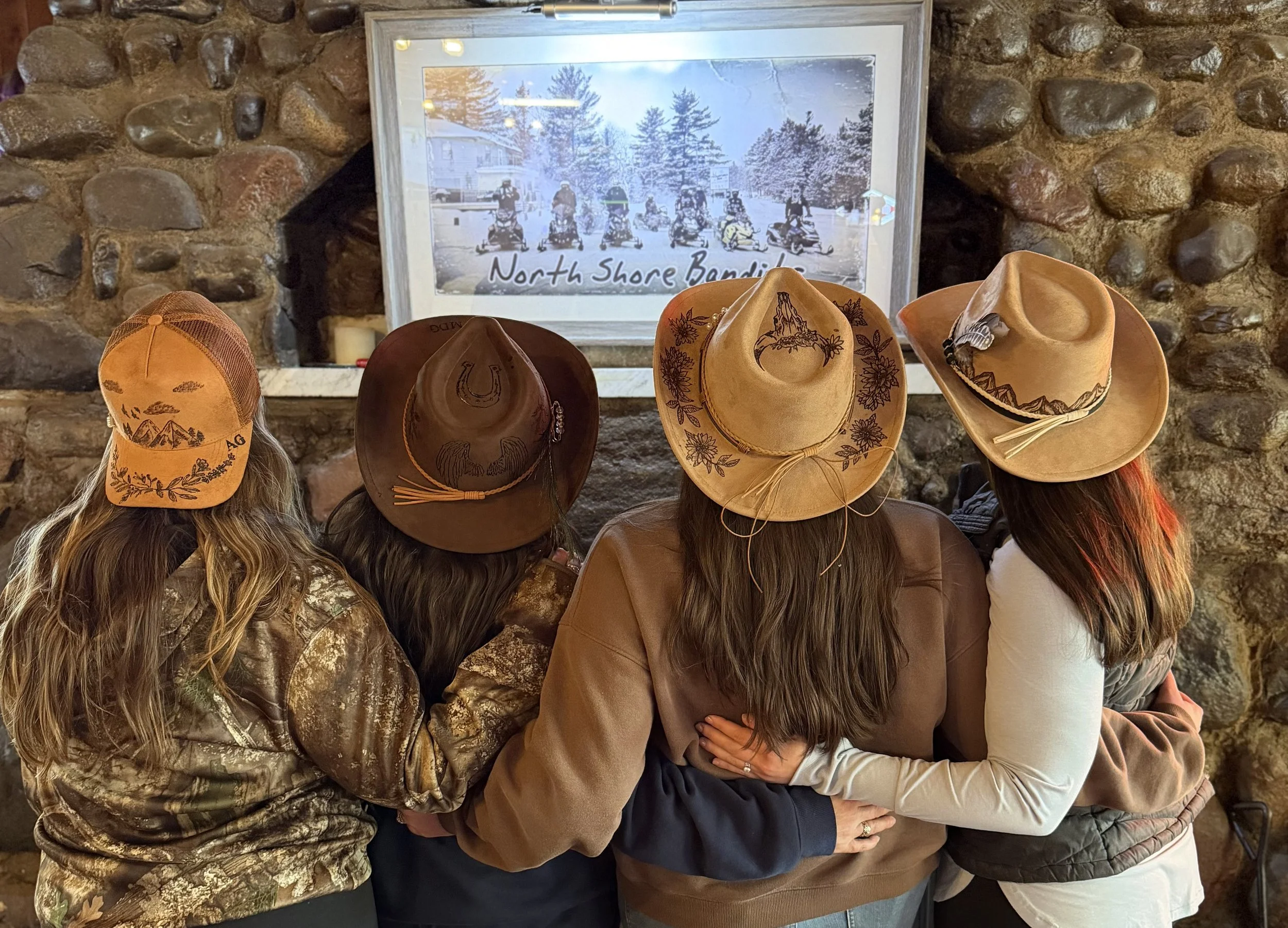 Four women with long hair wearing cowboy hats, standing in front of a stone wall and looking at a framed picture of snow-covered trees and ice skating people, with the text 'North Shore Bandits'.