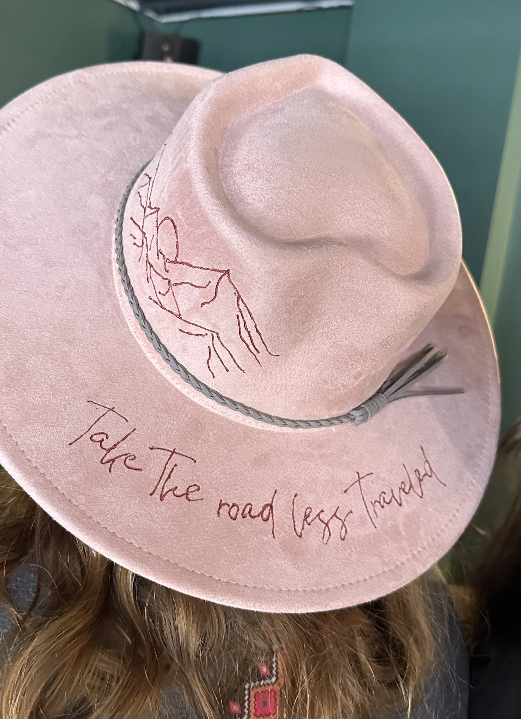 A pink hat with an illustration of a woman with long hair, along with handwritten script that reads, "To the road less traveled."