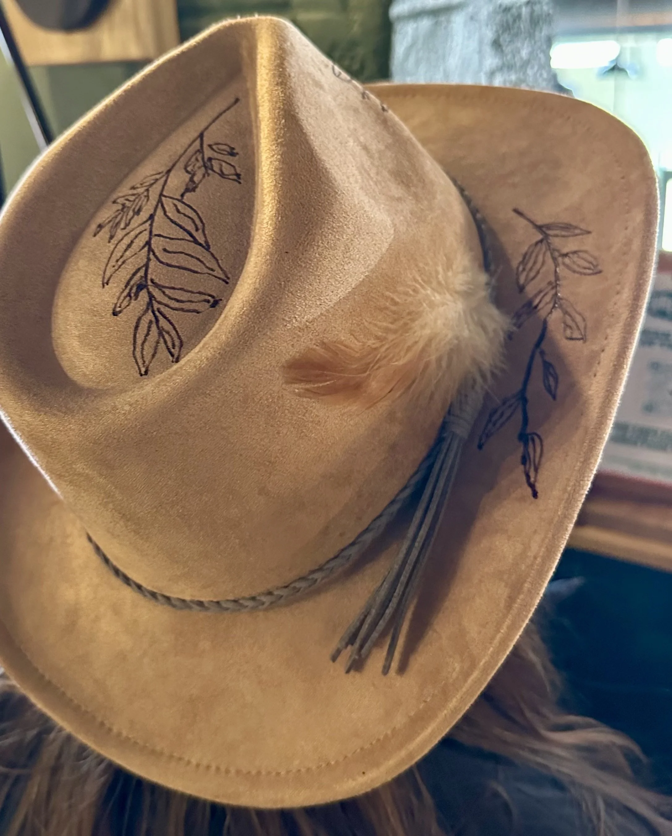 A tan cowboy hat with black embroidered leaves on the underside of the brim, resting on a surface with dark brown hair visible at the bottom.