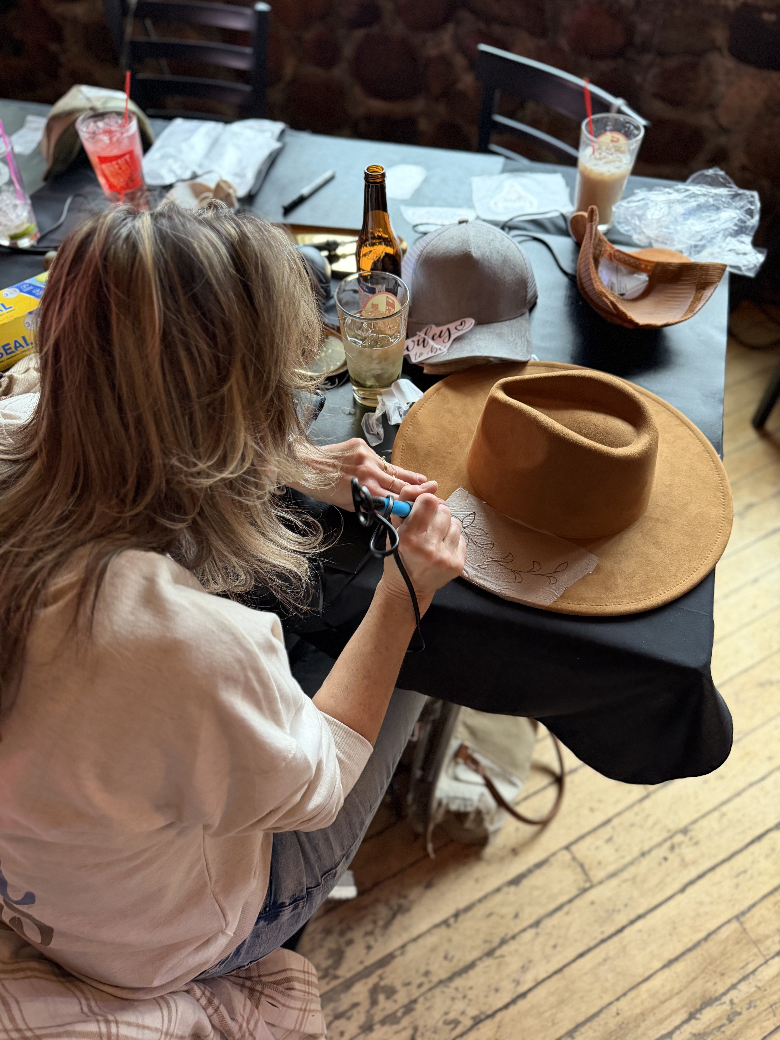 A person with blonde hair sits at a table with various drinks, hats, and personal items. The table has two partially consumed drinks in tall glasses, an empty beer bottle, and a gray cap with a pink tag. There are other objects like a brown hat, tiss