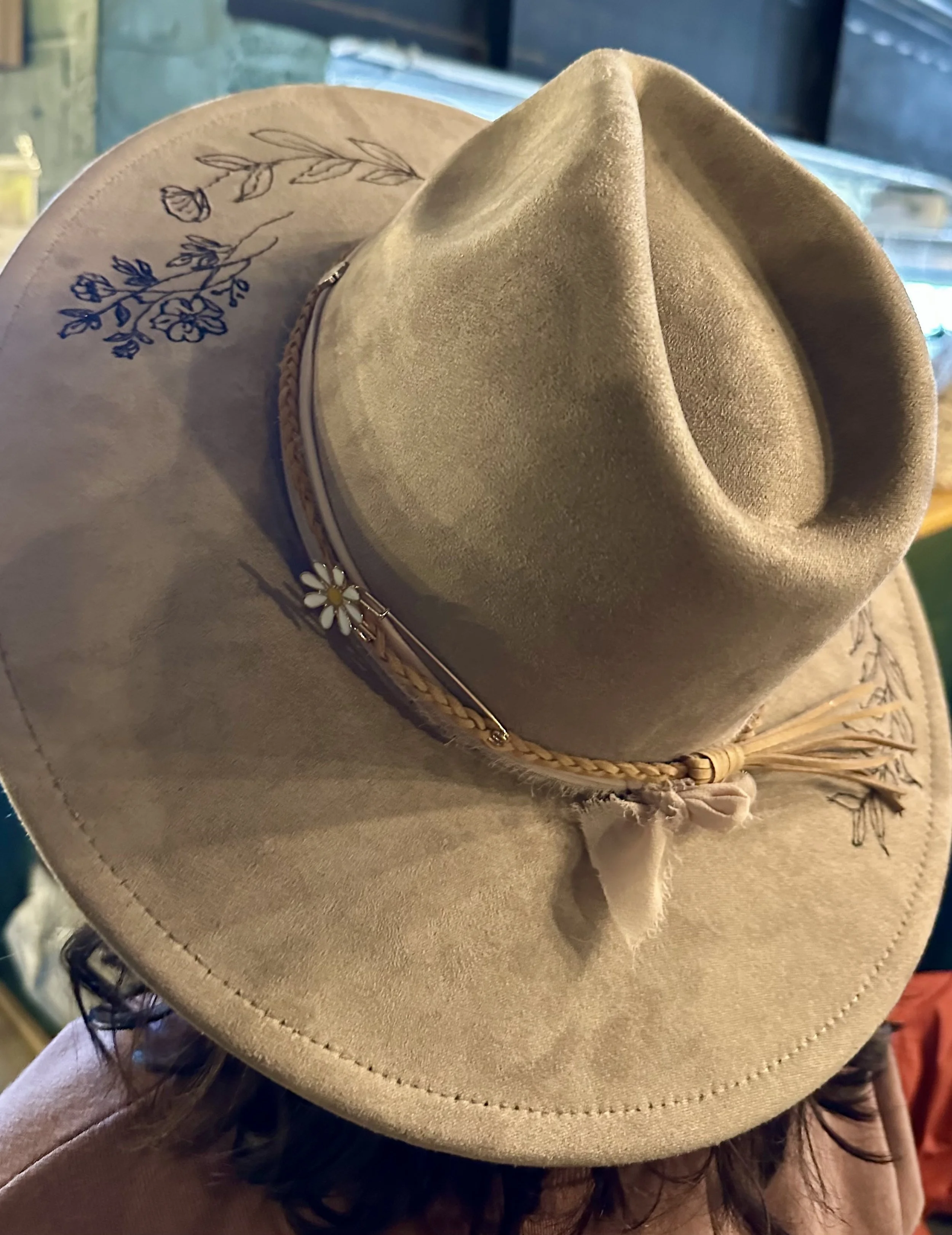 A beige cowboy hat with floral embroidery on the brim, set on top of a display of other hats.