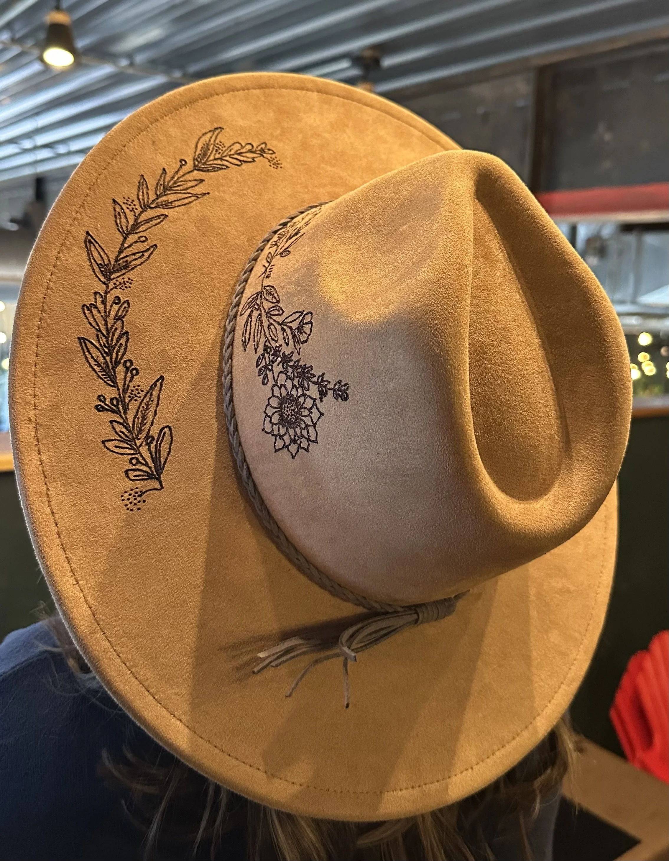 A tan wide-brimmed hat with black embroidered floral and leaf patterns on the brim, hanging on a person's head with long hair underneath, hung by a braided cord inside a rustic indoor setting.